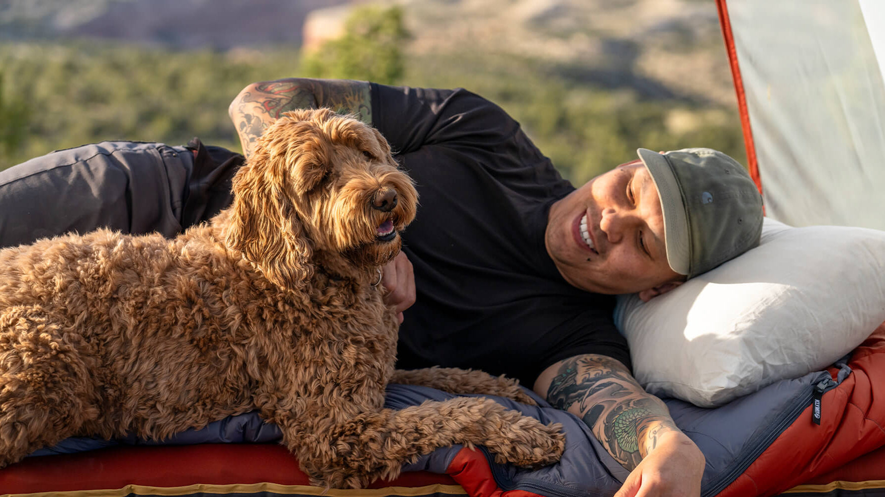 A person laing on a sleeping bag and pillow with a dog next to them.