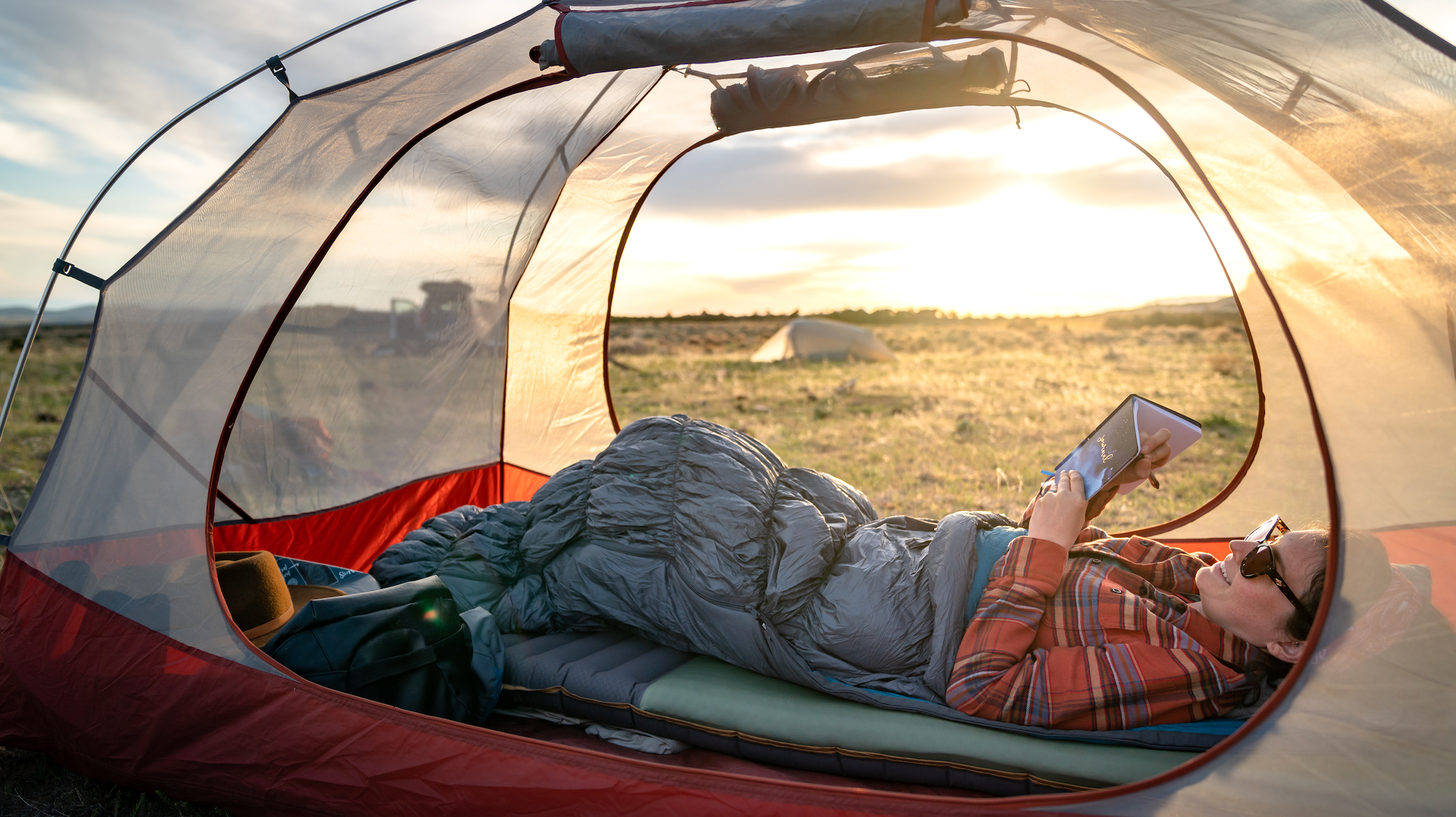 A woman laying in a sleeping bag inside a tent reading a book