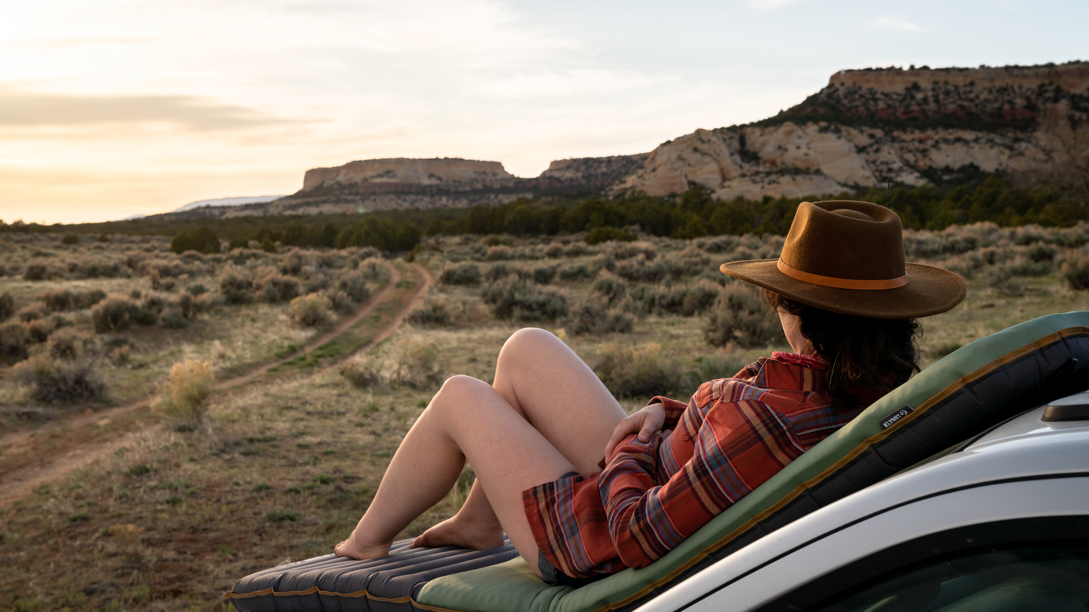 A woman resting on a sleeping pad on the hood of a car taking in the view of distant mountains