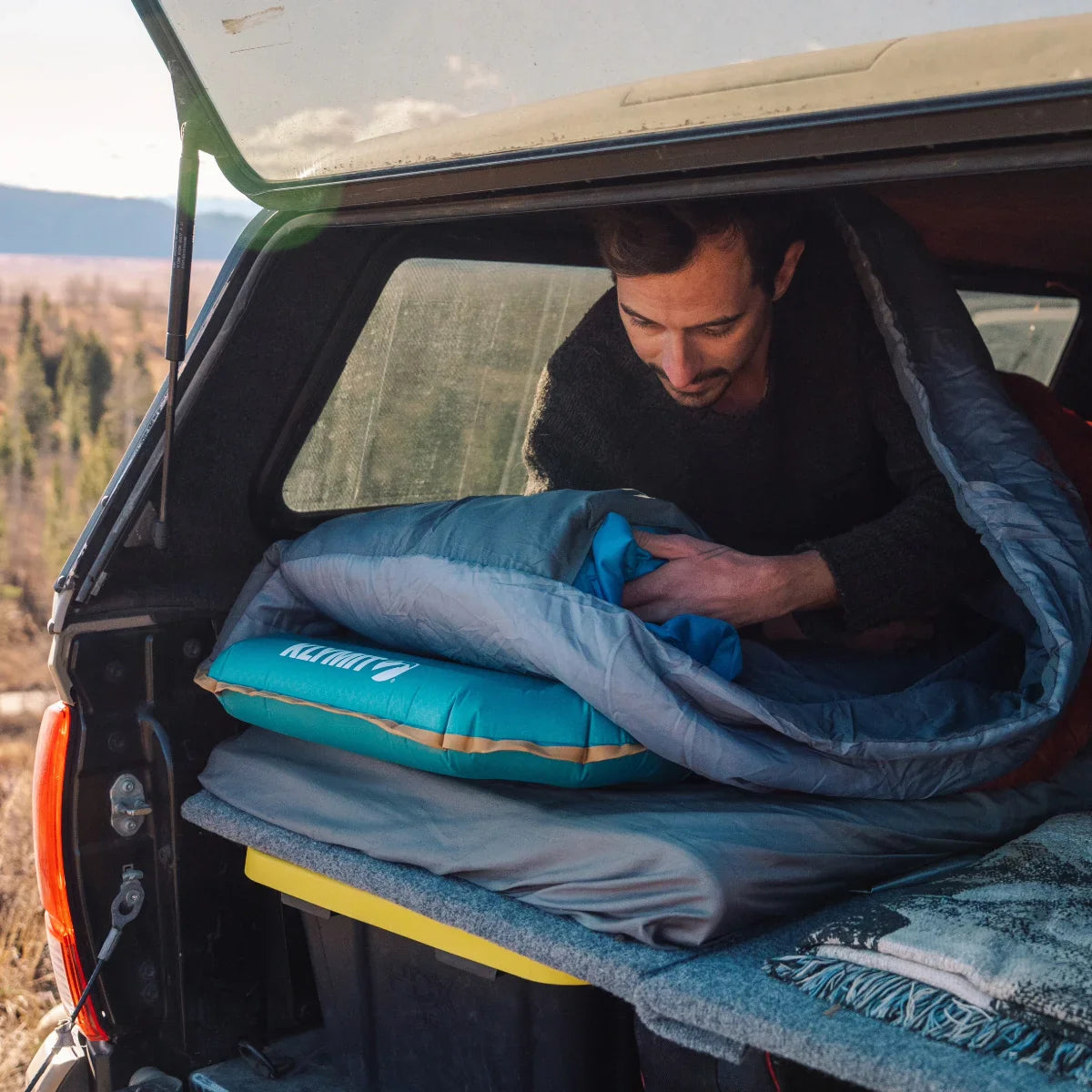 A man in a sleeping bag inside a covered pickup truck bed.
