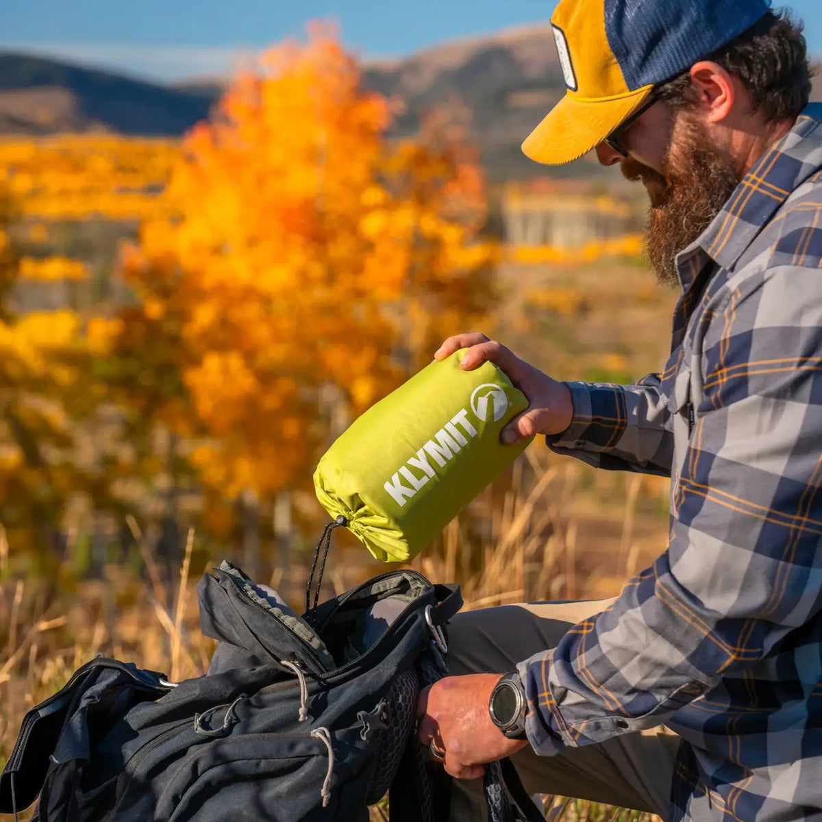 Bearded man in a flannel shirt packs a chartreuse Klymit Static V Summit sleeping pad in its stuff sack into a backpack.