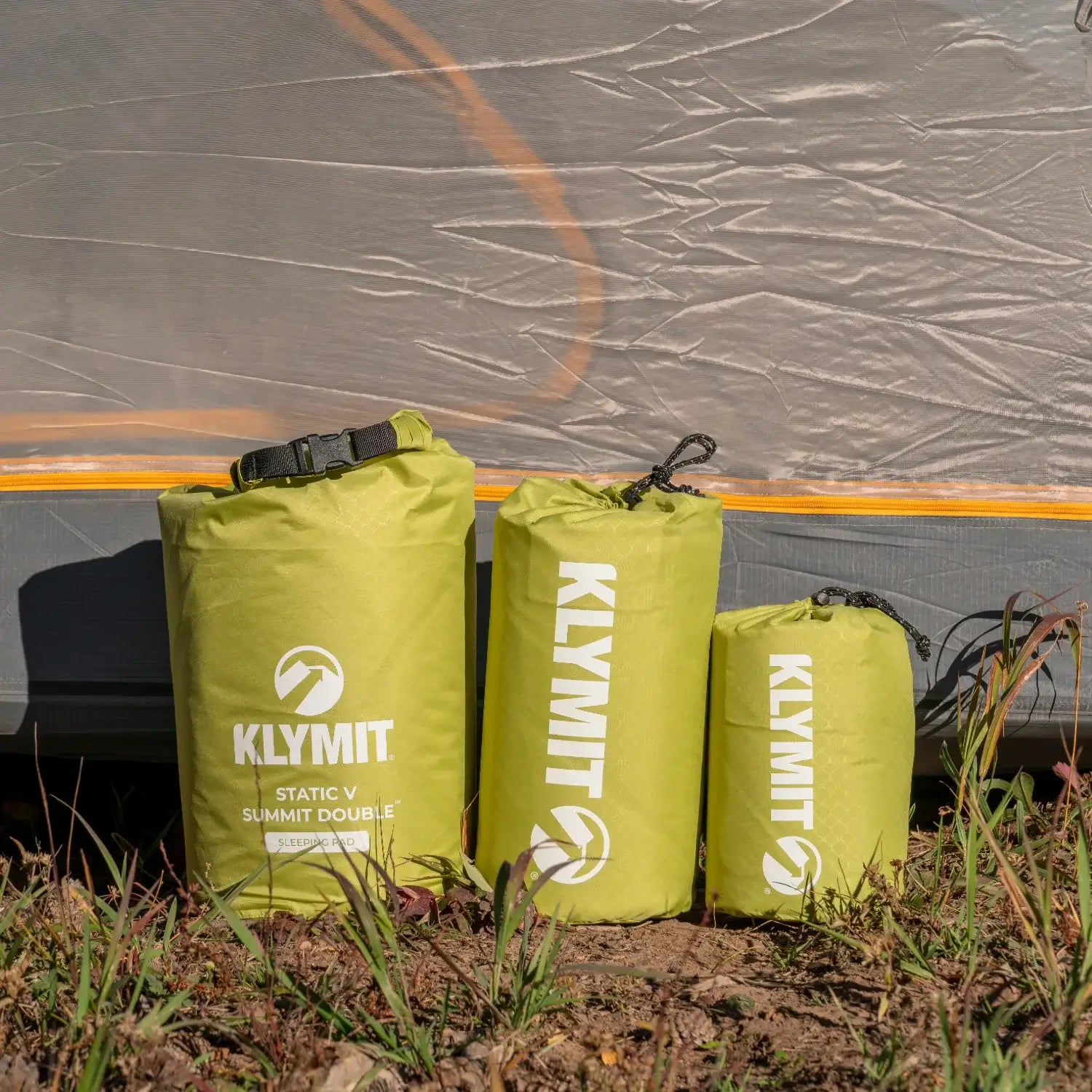 Three sizes of Klymit Static V Summit sleeping pads in chartreuse stuff sacks lined up next to a tent at an outdoor campsite.