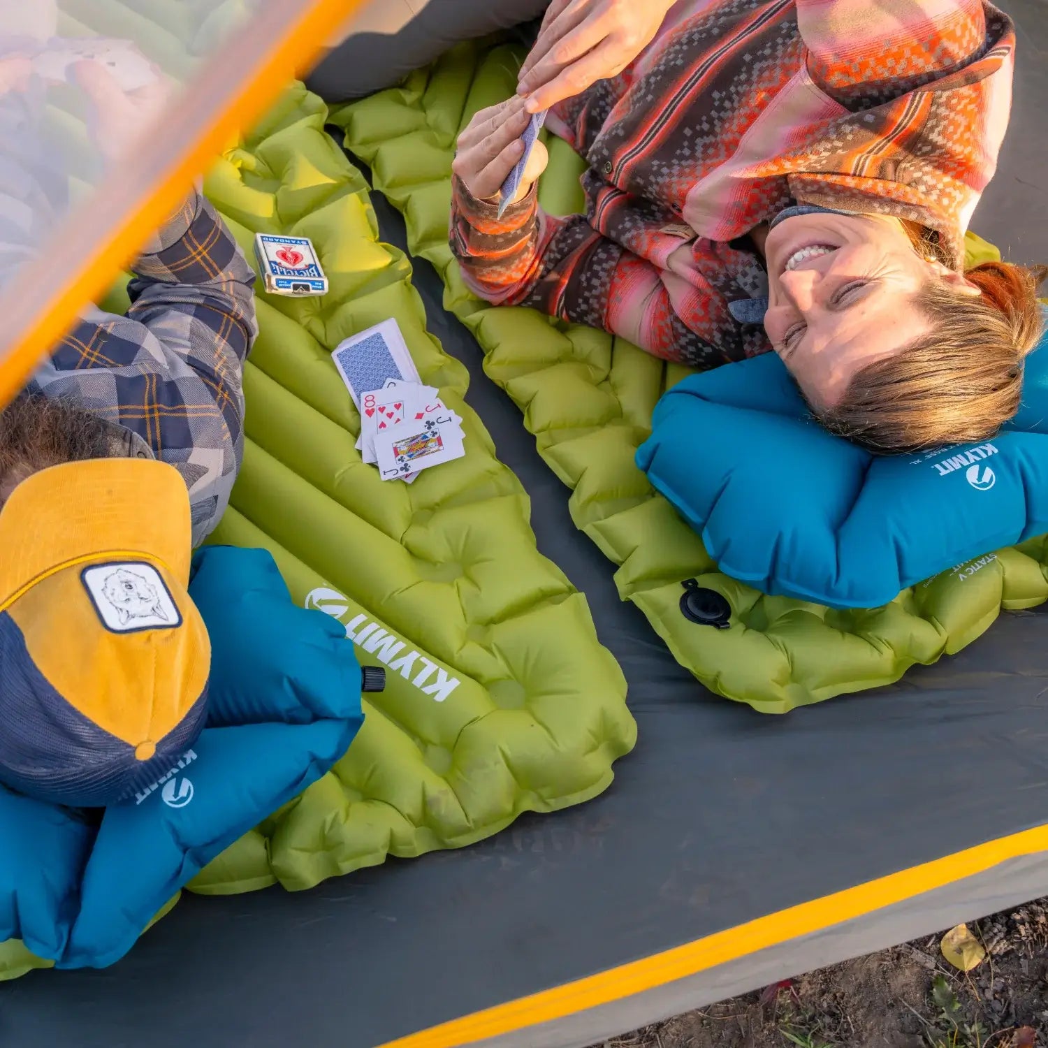 Overhead view of a couple relaxing on chartreuse Klymit Static V Summit sleeping pads and a deck of playing cards spread between them inside a tent.