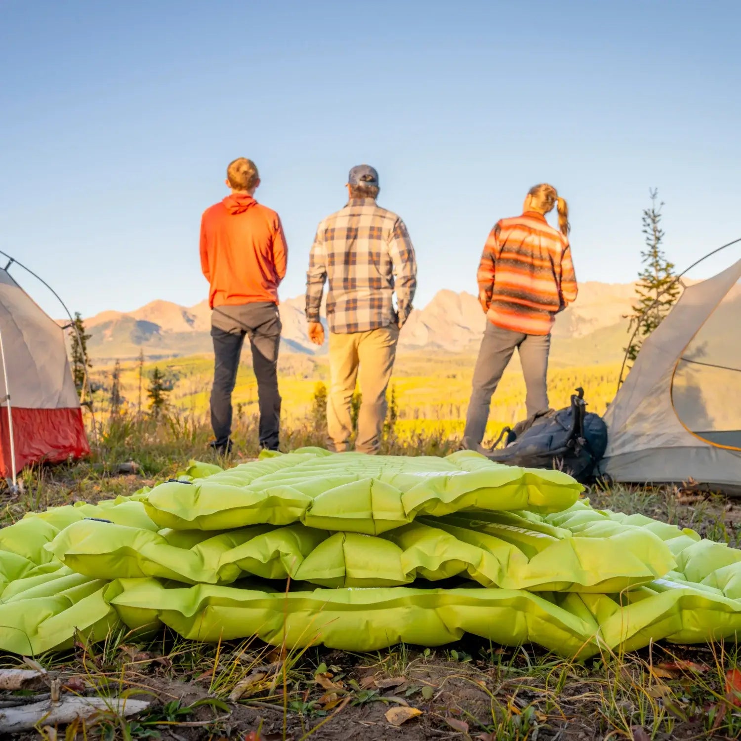 Three campers standing outside their tents looking out over a mountain valley at sunset, with stacked chartreuse Klymit Static V Summit sleeping pads in the foreground.