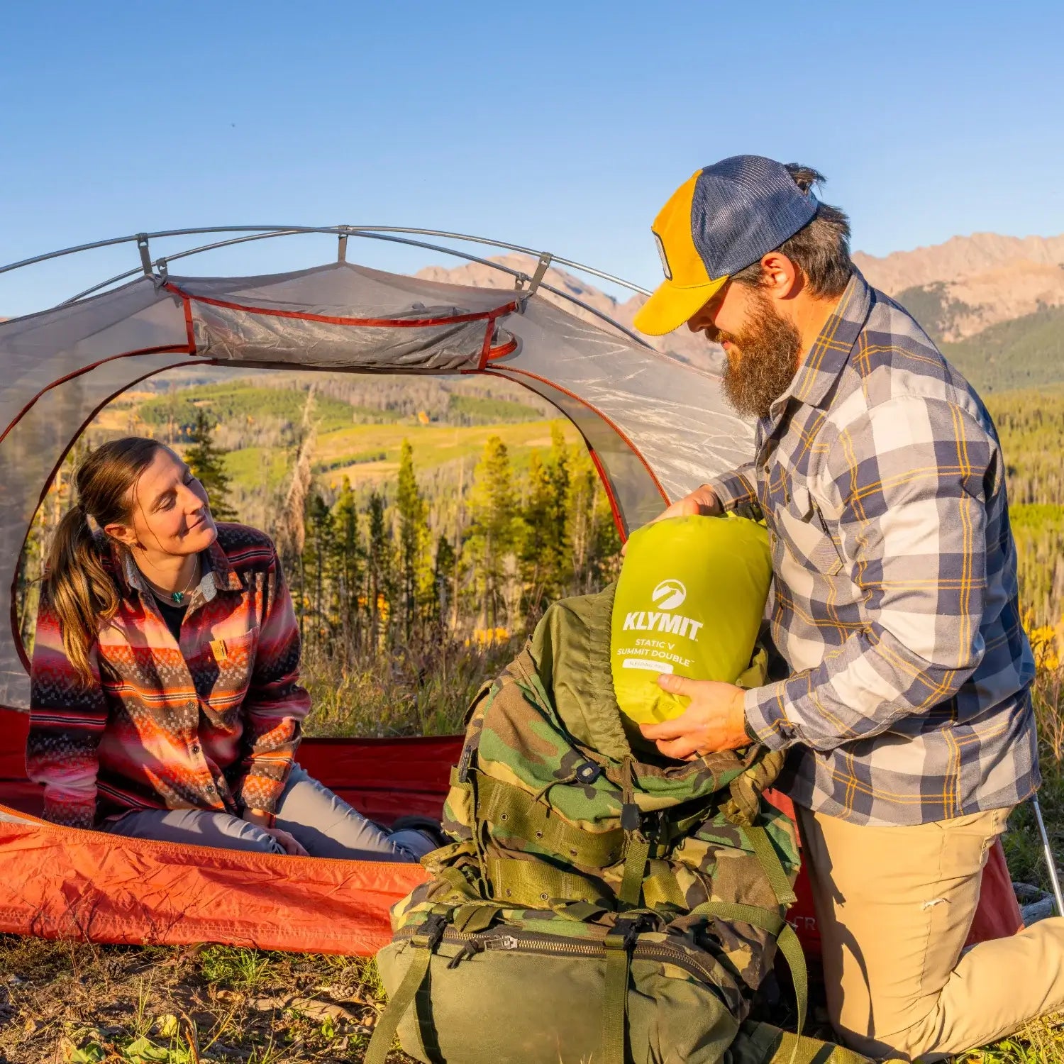 Man packing a chartreuse Klymit Static V Summit Double sleeping pad into a backpack while a woman watches from inside a tent overlooking a mountain valley.