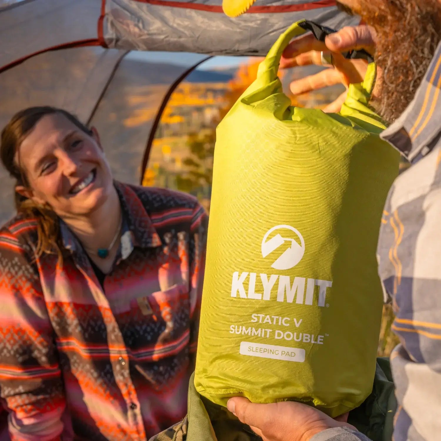 Close-up of a man holding a chartreuse Klymit Static V Summit Double sleeping pad in its stuff sack, with a smiling woman in plaid visible in the background.