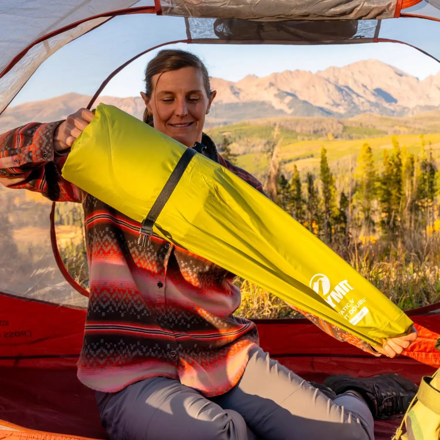 Woman inside a tent unpacking a chartreuse Klymit Static V Summit Double sleeping pad from its stuff sack, with mountain views visible through the tent door.