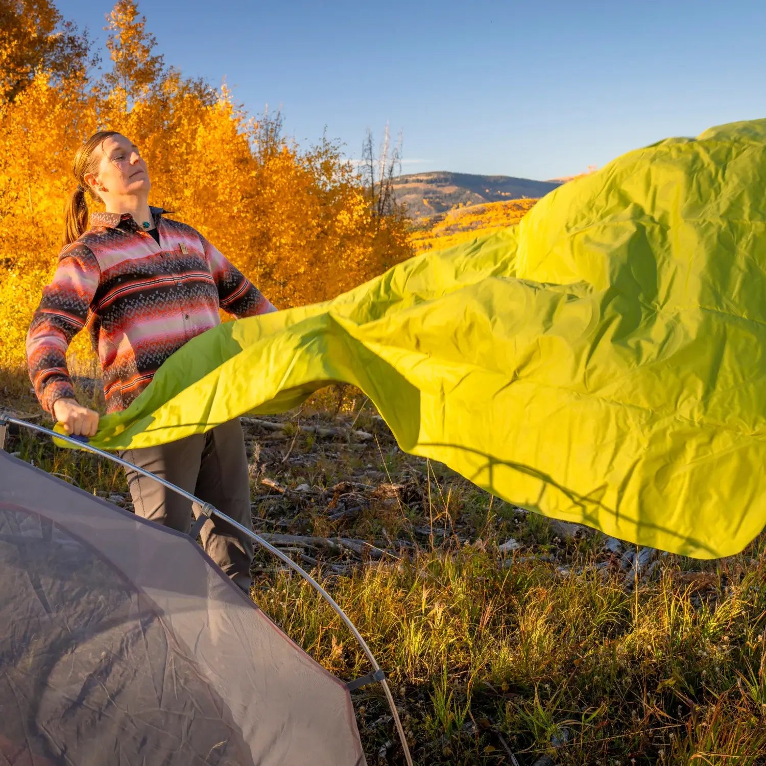 Woman unfolding a chartreuse Klymit Static V Summit Double sleeping pad to inflate it outside a tent.