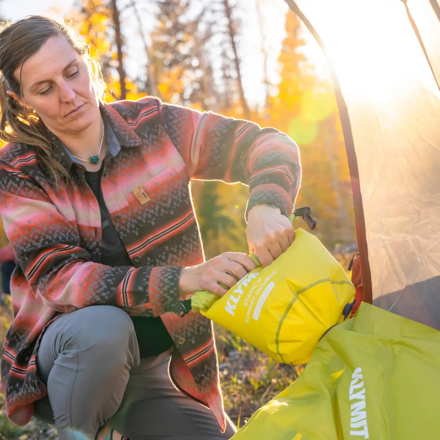 Woman rolling up a chartreuse Klymit Static V Summit Double sleeping pad into its stuff sack outside a tent in golden afternoon light.