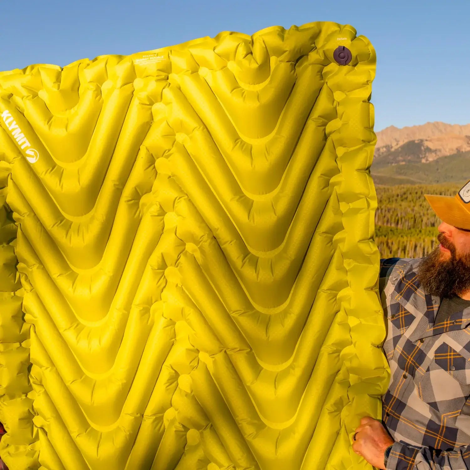 Man holding up an inflated chartreuse Klymit Static V Summit Double sleeping pad showing its V-chamber design, with mountains in the background.