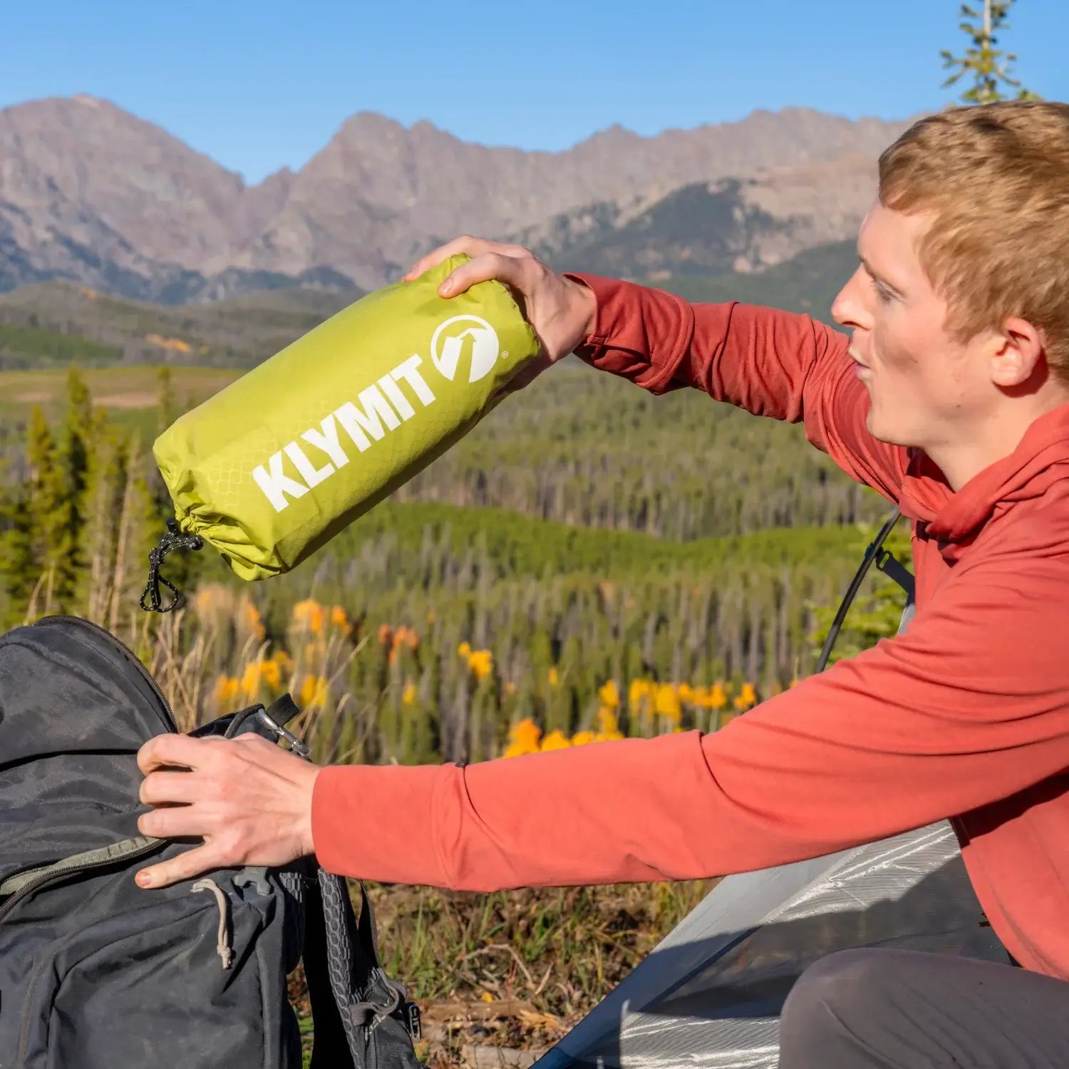 Man pulling a Klymit Static V Summit sleeping pad in chartreuse stuff sack from a backpack with mountains and pine trees in the background.