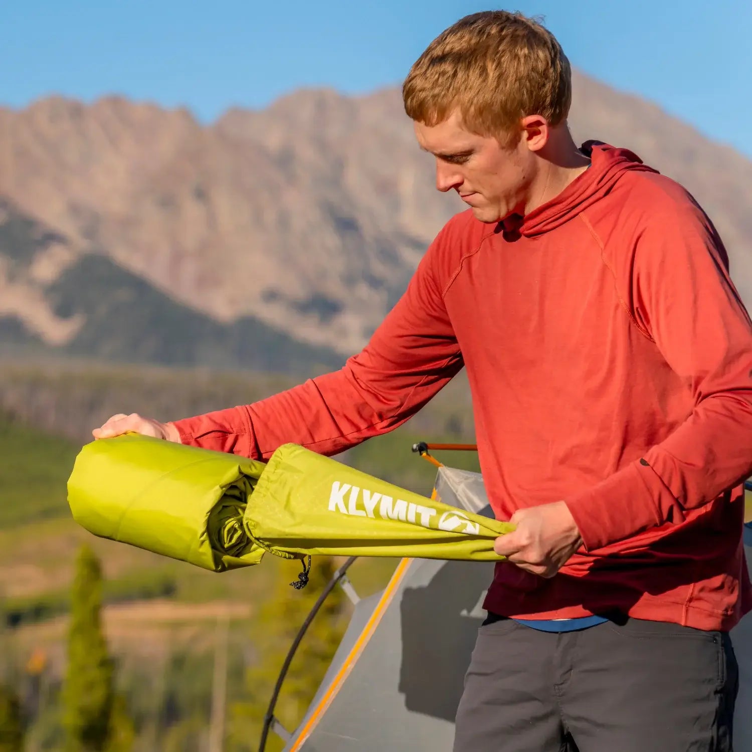 Man removing a chartreuse Klymit Static V Summit sleeping pad from its stuff sack outside a tent with mountain scenery behind.