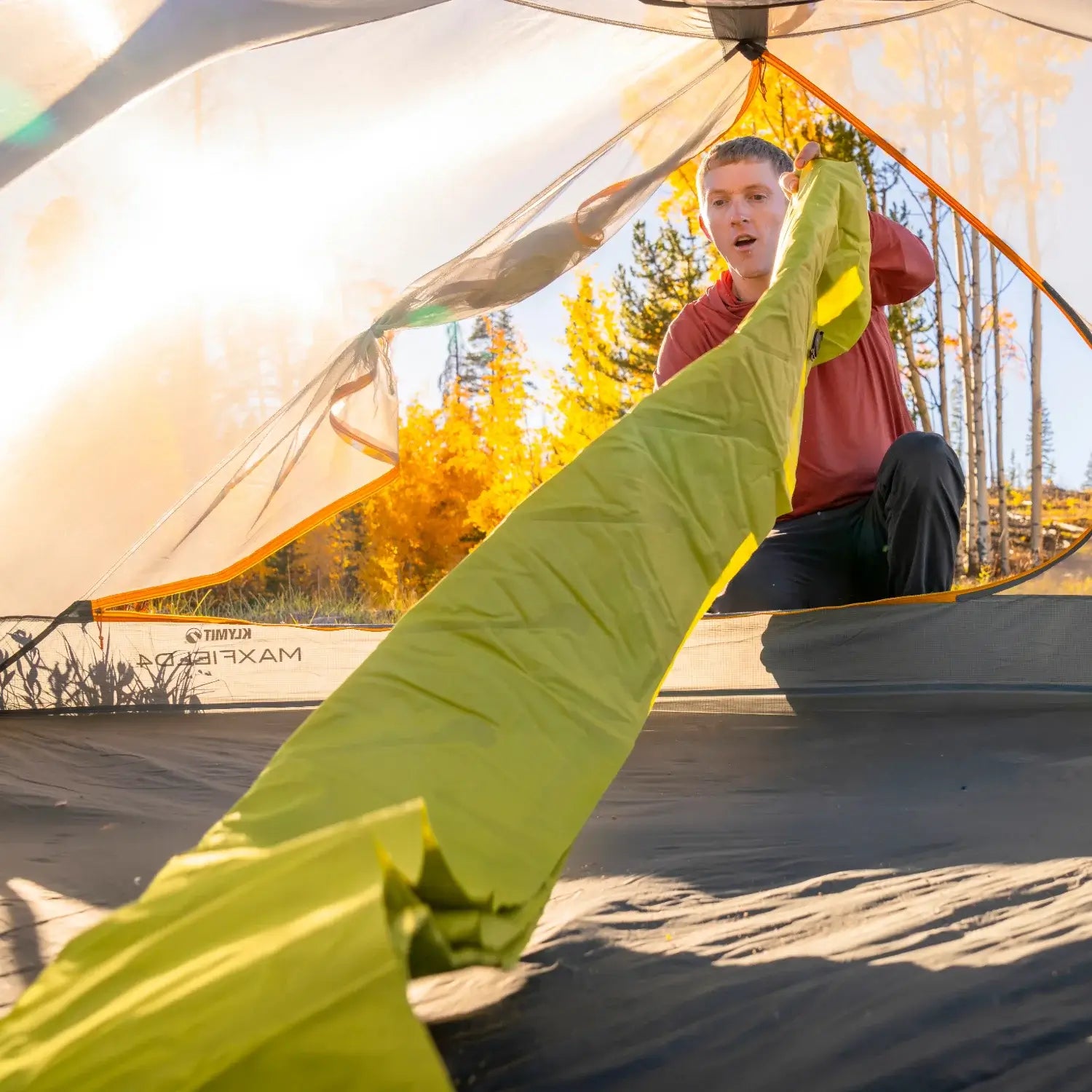 Man unrolling a chartreuse Klymit Static V Summit sleeping pad inside a tent with golden autumn aspens visible through the doorway.