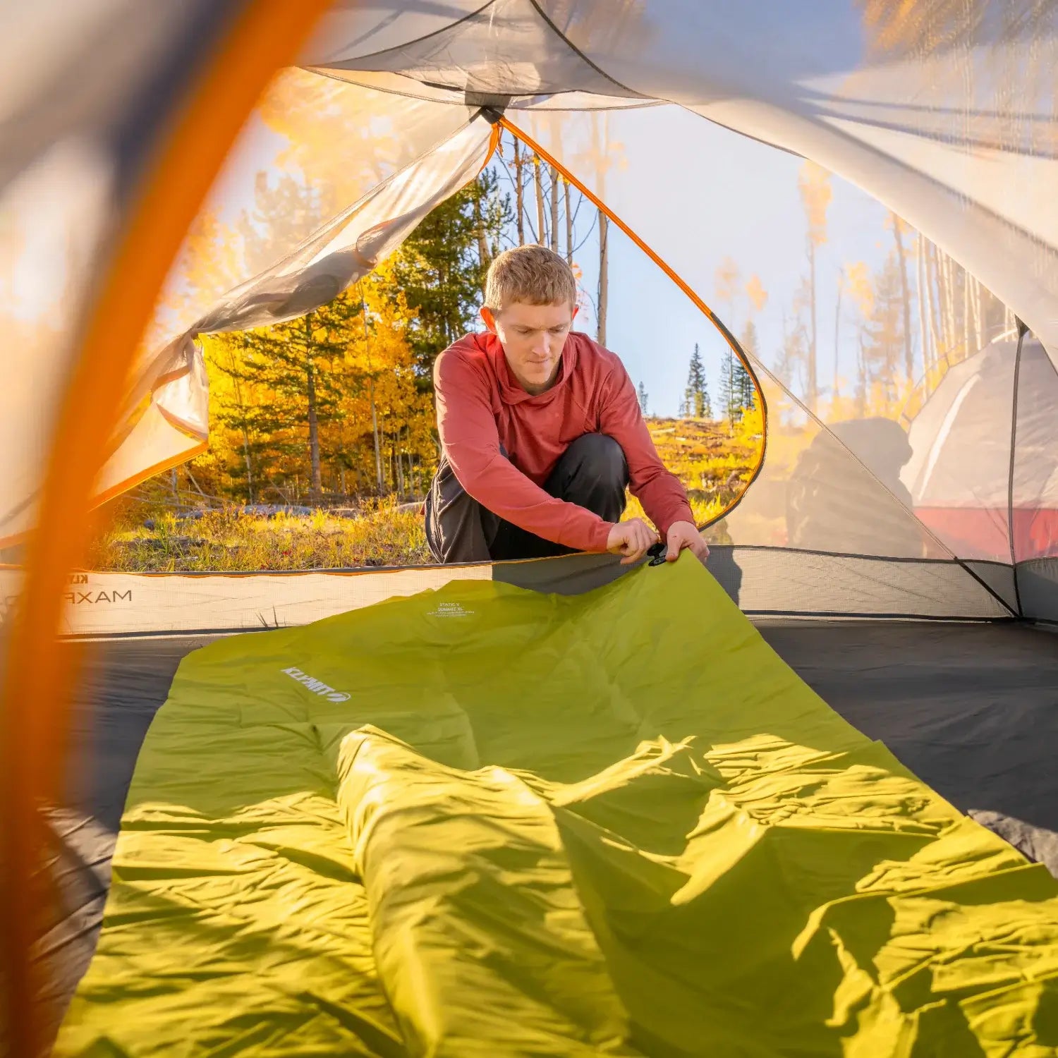 Man setting up a chartreuse Klymit Static V Summit sleeping pad inside a tent at a sunlit fall campsite.