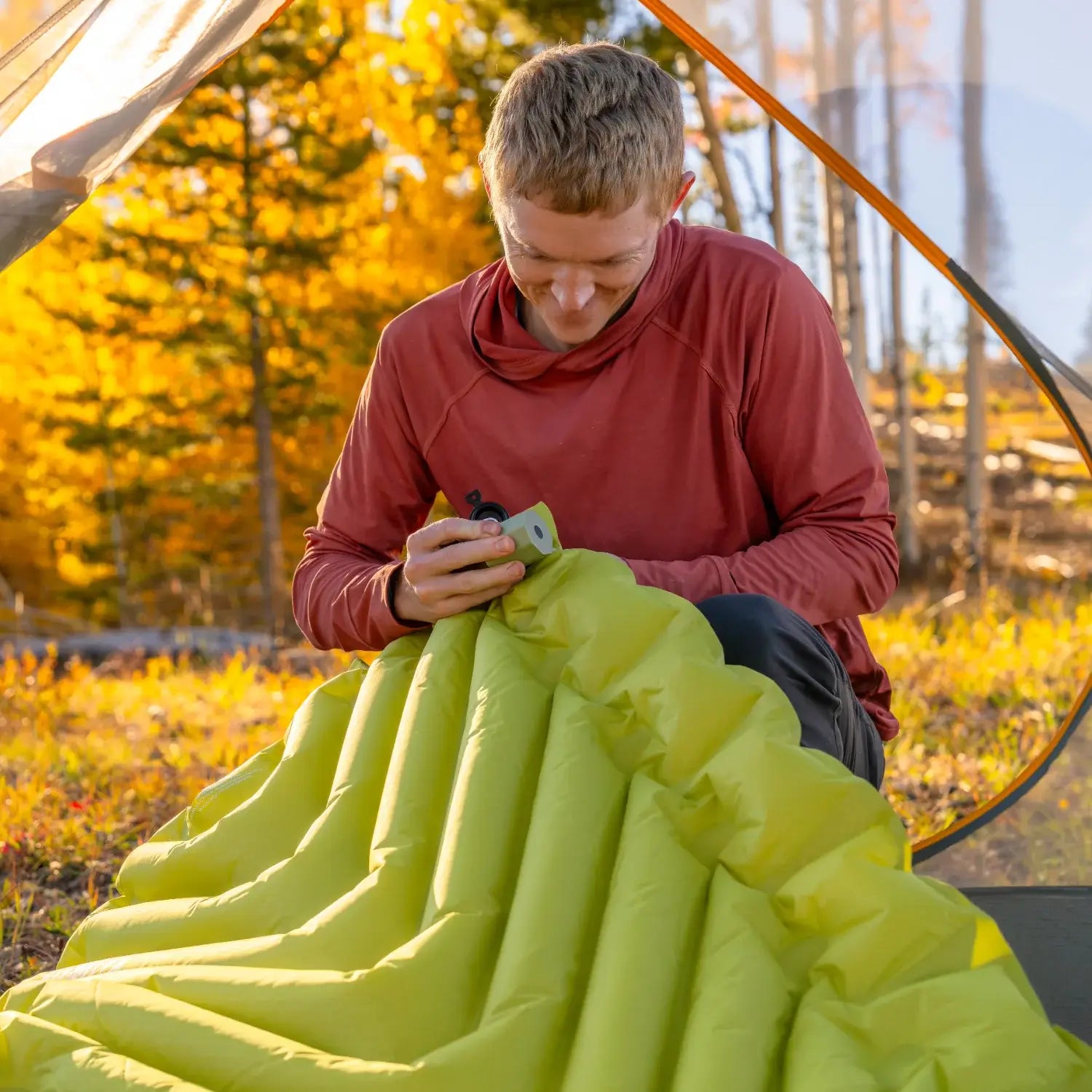 Man closing the valve on an inflated chartreuse Klymit Static V Summit sleeping pad inside a tent surrounded by autumn foliage.