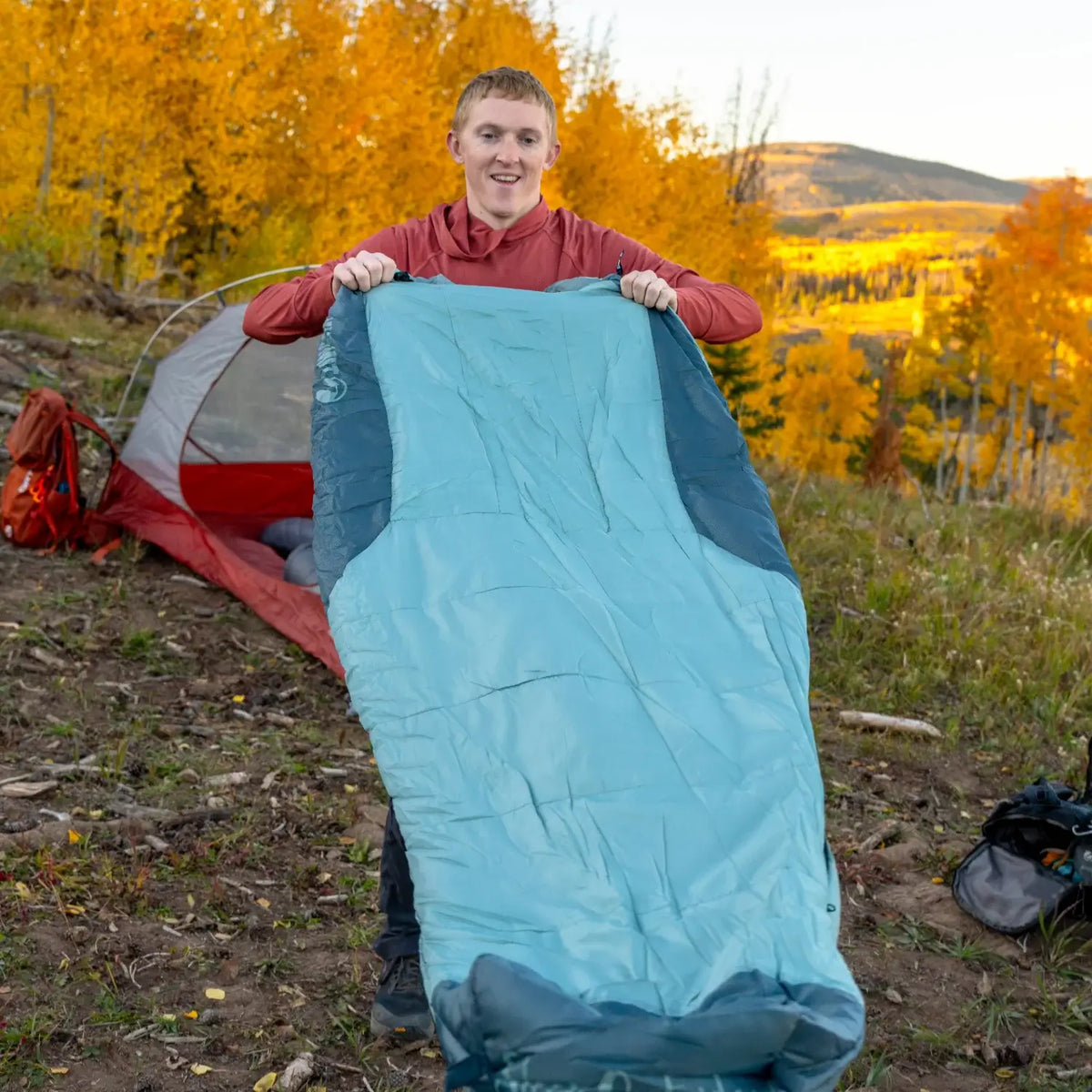 Man holding open a Klymit Wild Aspen 20 Large sleeping bag at a fall campsite with mountain views.