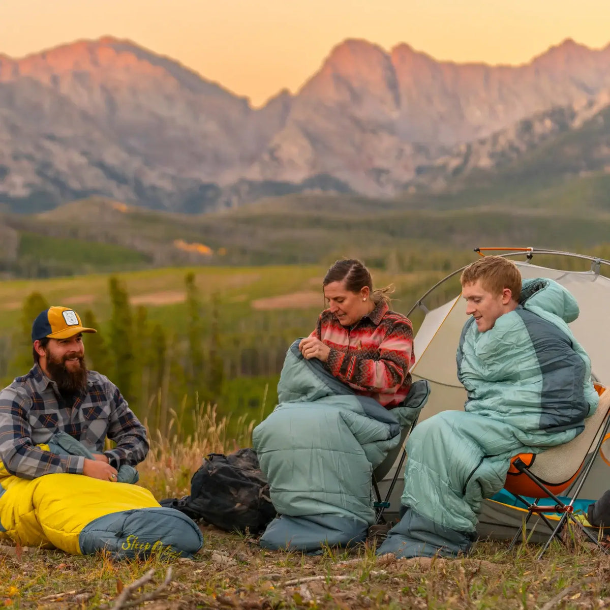 all-groups Three campers in Klymit Wild Aspen sleeping bags at sunset with a mountain backdrop.