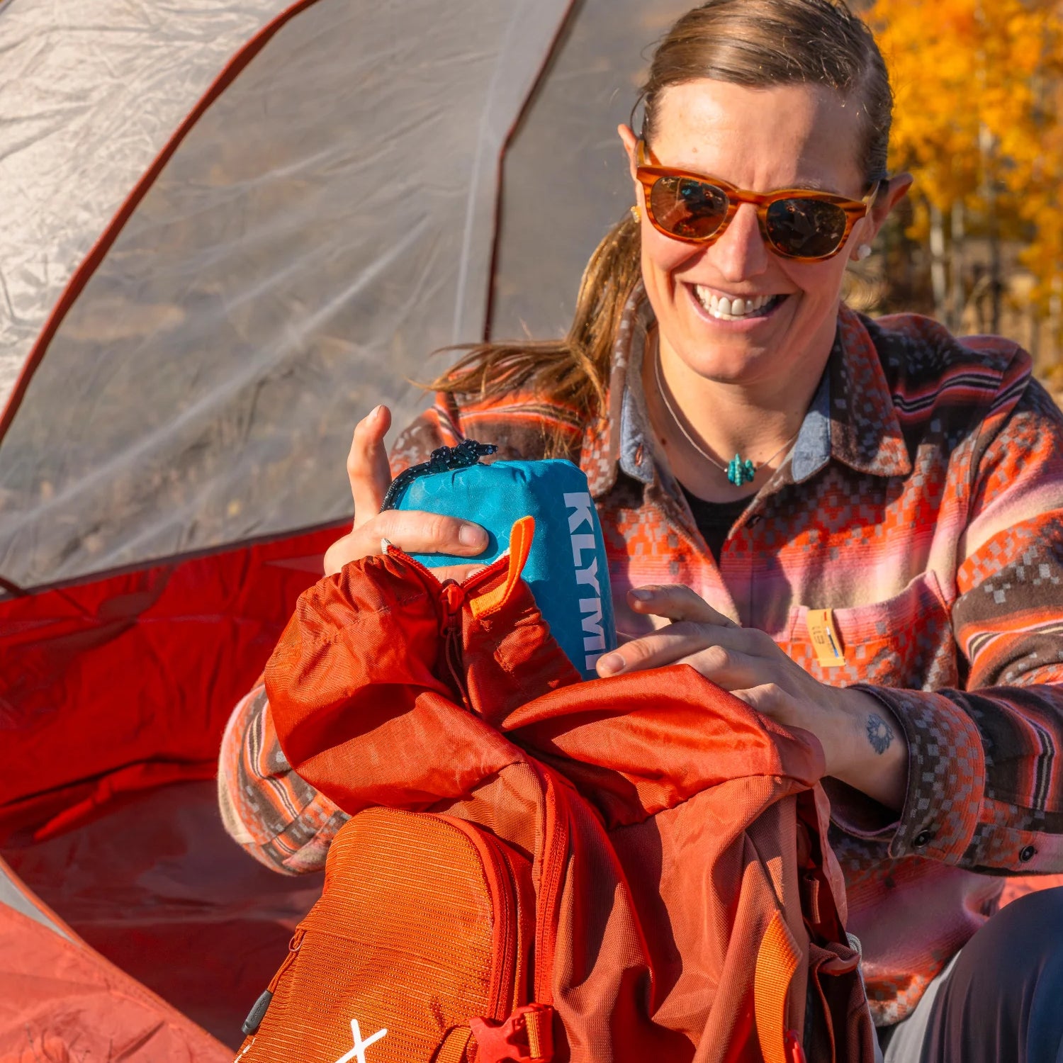 Smiling woman pulling a Klymit sleeping pad stuff sack out of her backpack outside a tent surrounded by golden autumn aspens.