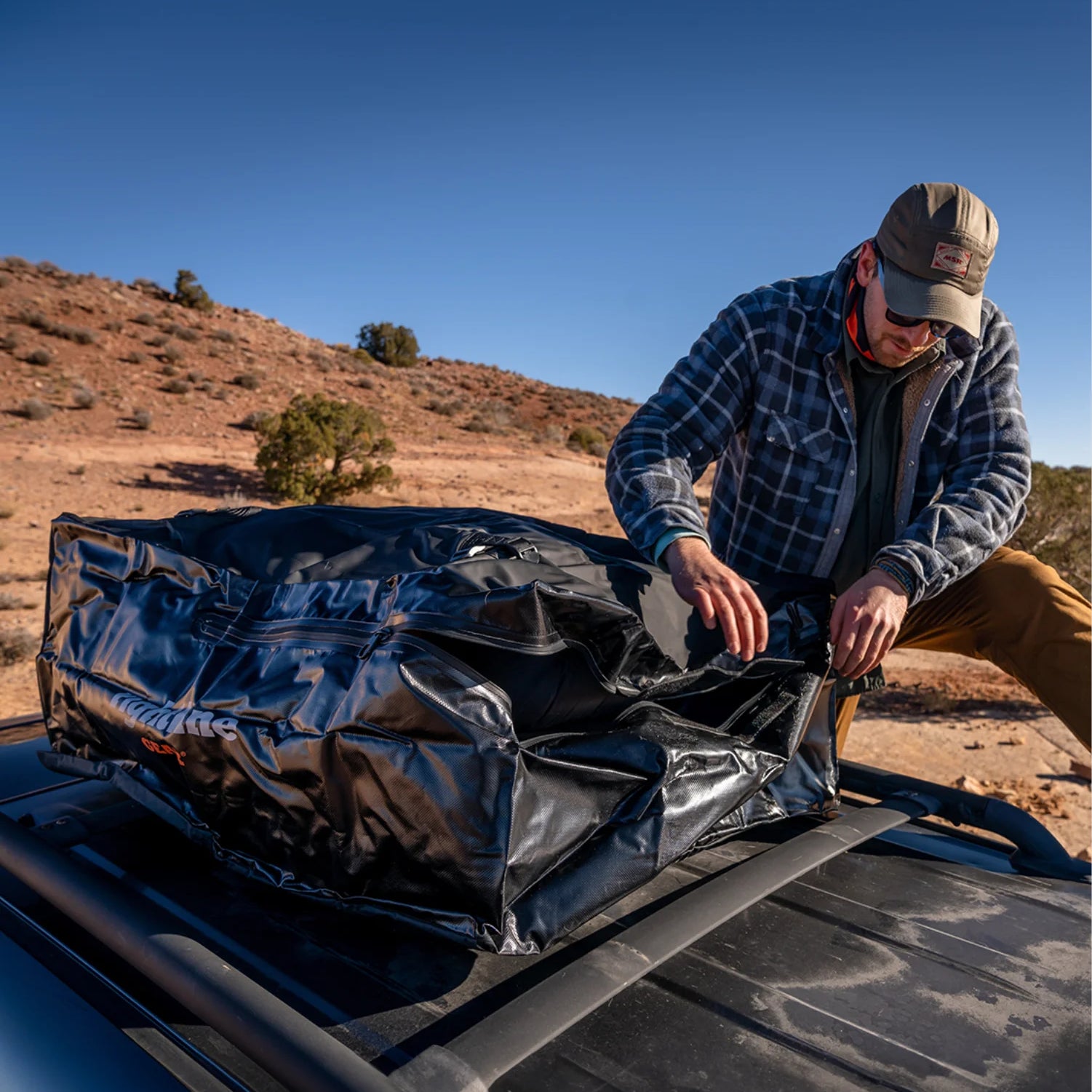 Man loading a Rightline gear waterproof rooftop cargo carrier onto a vehicle roof rack at a remote desert overlanding site.