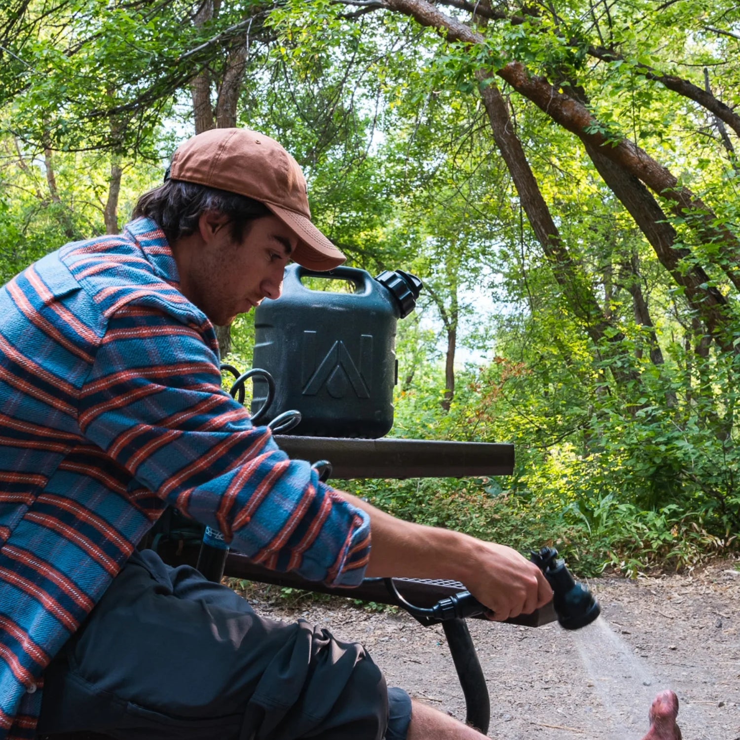Man rinsing his foot using a WaterPORT GoSpout portable water tank with spray nozzle at a wooded campsite.