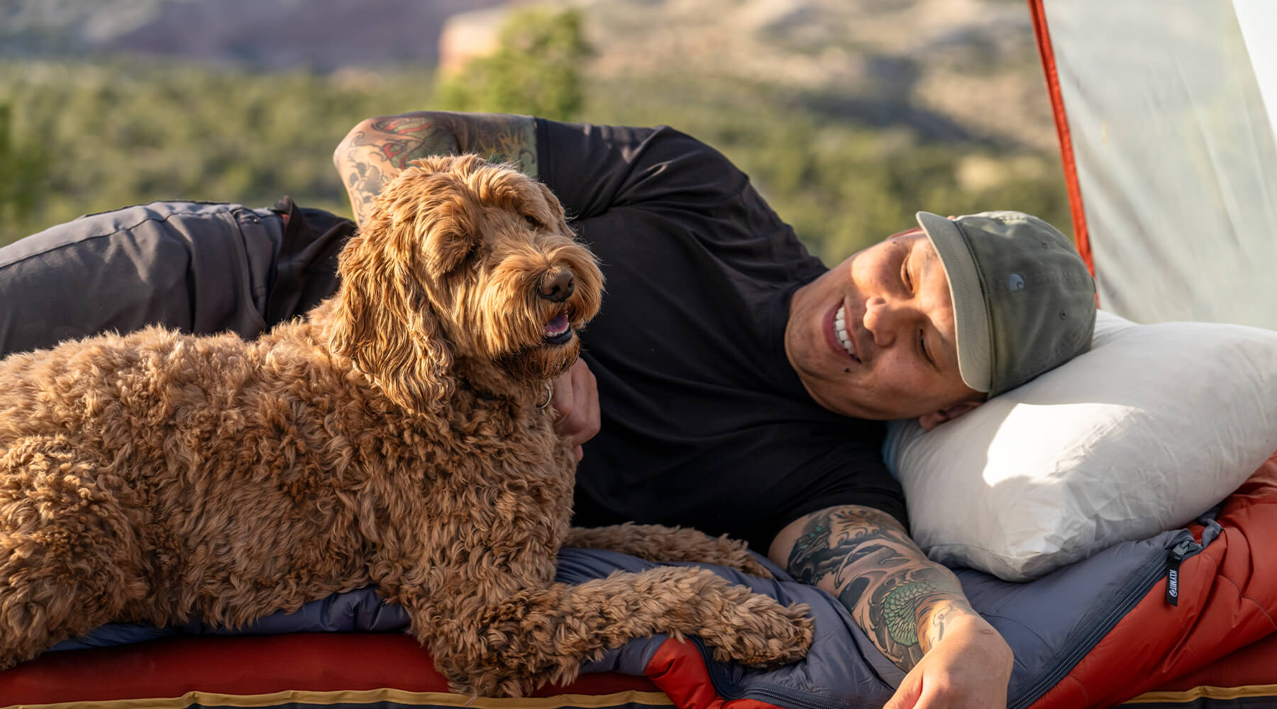 A person laing on a sleeping bag and pillow with a dog next to them.