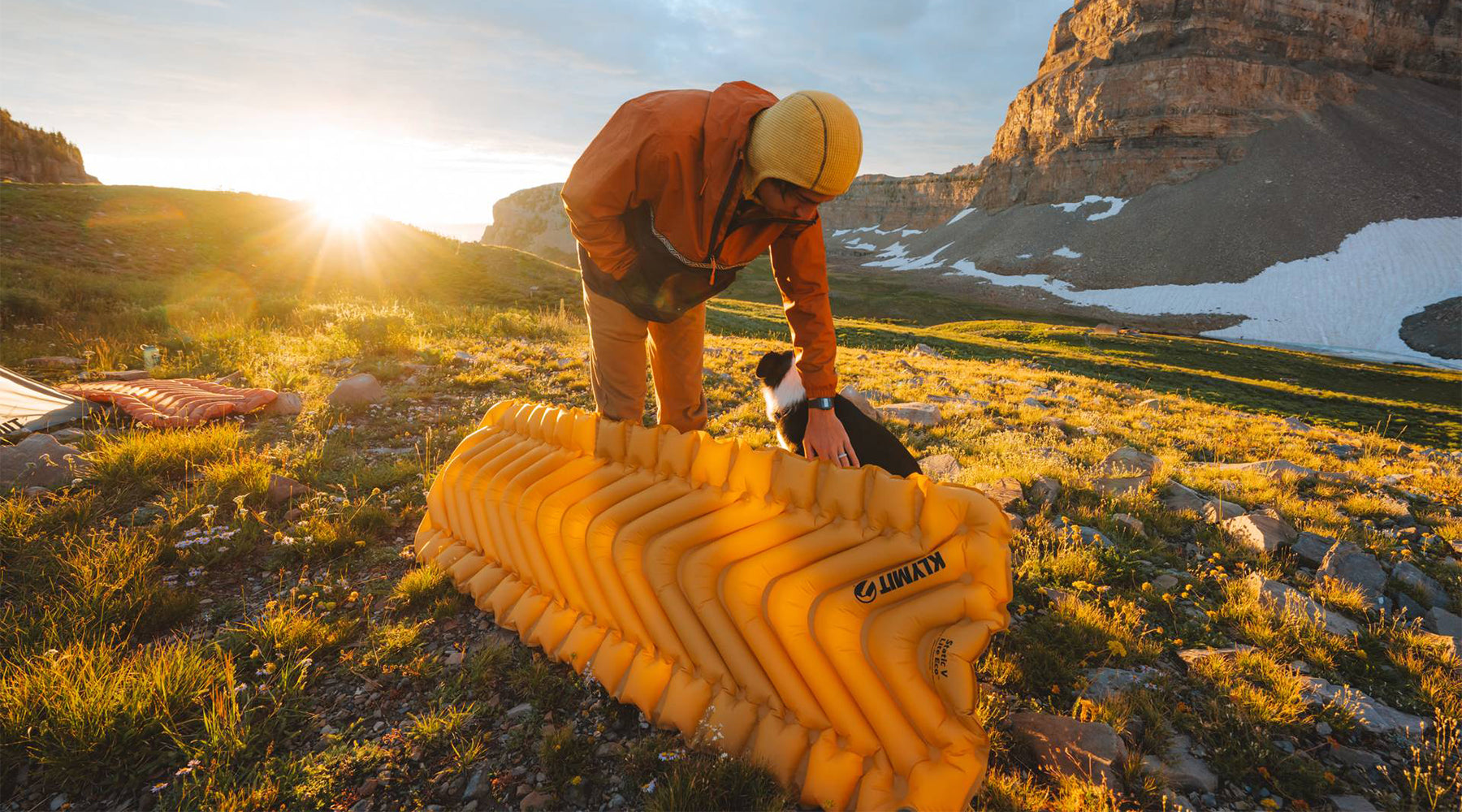 A person petting a dog with a Klymit V Sleeping pad propped against their leg.
