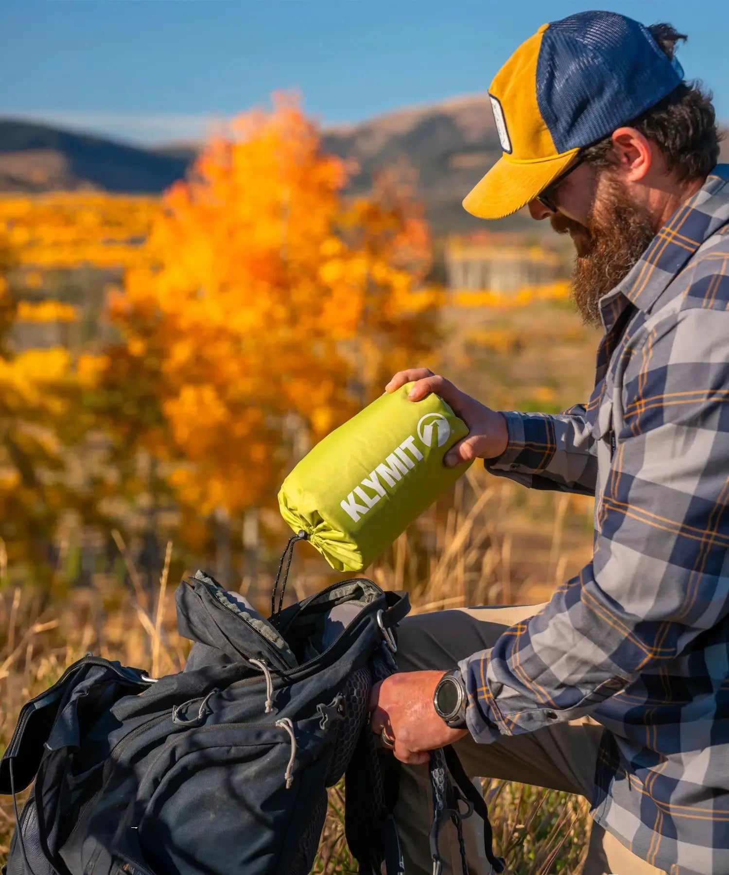 Bearded man in a flannel shirt packs a chartreuse Klymit Static V Summit sleeping pad in its stuff sack into a backpack.