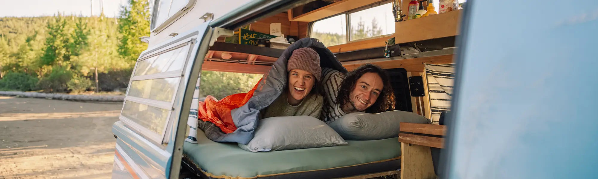 Smiling couple lie on a green Klymit Klymaloft Summit sleeping pad with gray Drift camp pillows and an orange Bozeman 35 sleeping bag inside the open side door of a vintage camper van.