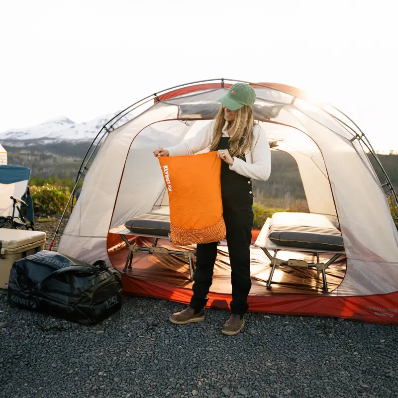 A woman wearing a green cap, white sweater, and black overalls holds an orange Klymit Drift Pillow in front of a large, open camping tent with two cots inside. The sun is setting behind her, casting a golden glow over the mountain landscape in the background.