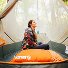 A woman in a colorful quilted jacket sits inside a tent, looking up peacefully. In front of her is an orange Klymit Drift Pillow resting on the tent floor, with trees and soft natural light visible through the mesh walls.