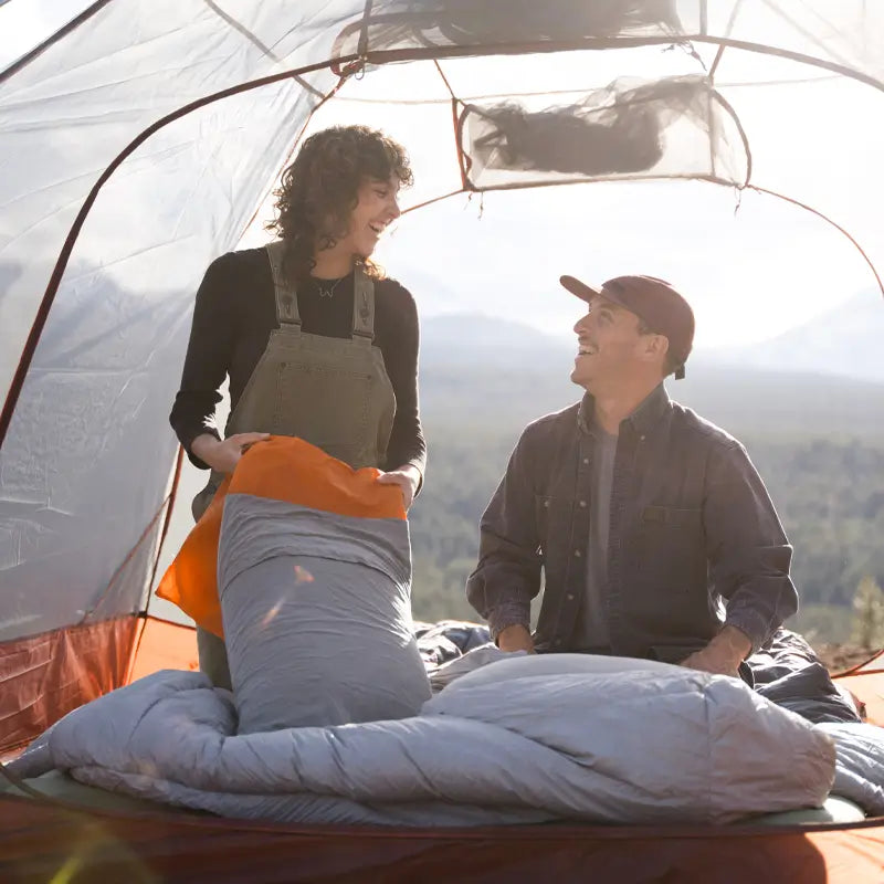 A smiling couple inside a camping tent, with the woman holding a gray sleeping bag with an orange Klymit Drift Pillow on top. The man, wearing a cap and denim shirt, looks up at her as they enjoy the mountain view in the background.