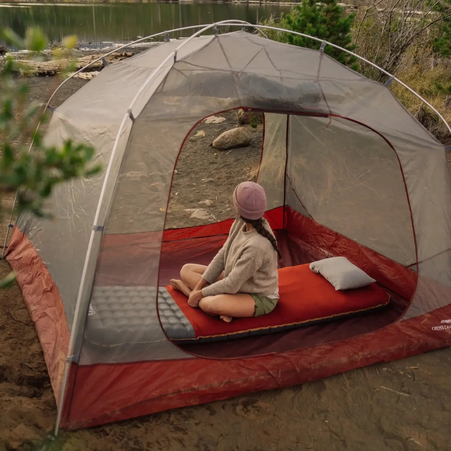 Woman sitting on a Klymit Insulated Klymaloft Peak XL sleeping pad inside a Klymit Cross Canyon tent by a lake, rear view.