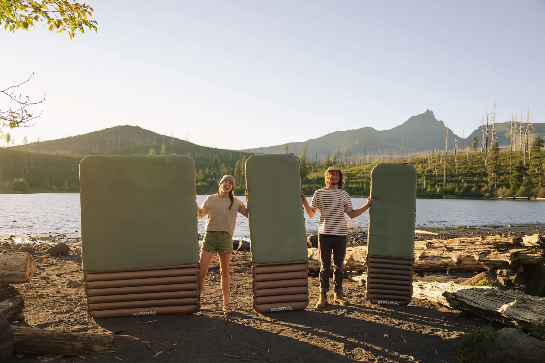 Couple holding up Klymit Klymaloft Summit foam sleeping pads at a sunlit lakeside campsite with mountain views in the background.