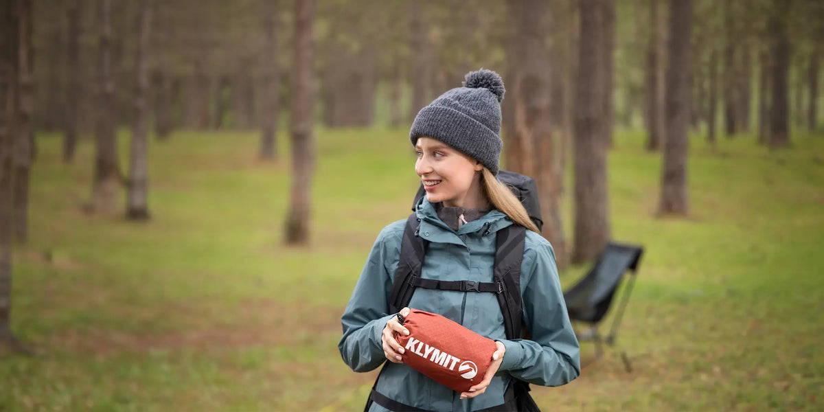 Woman in a beanie and rain jacket holding a red rock-colored Klymit sleeping pad packed in its stuff sack beside a tent.