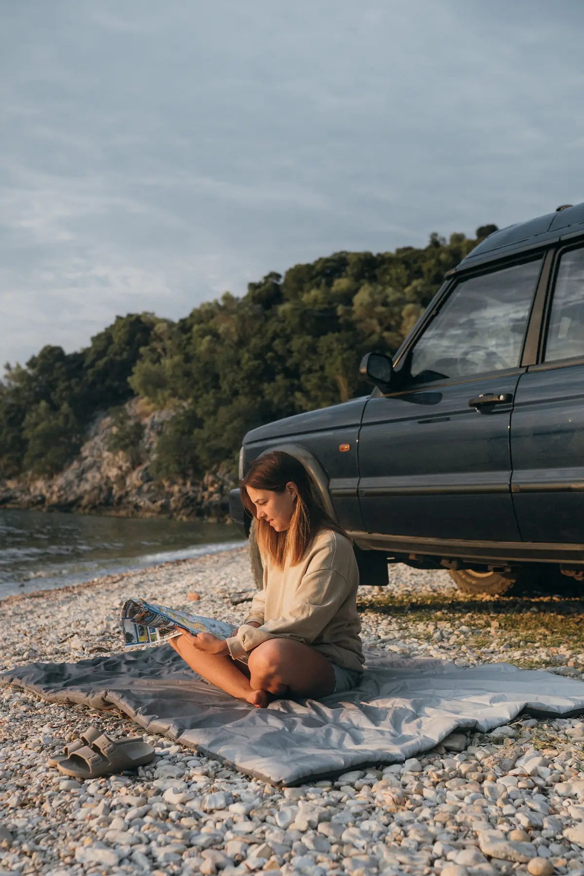A woman sitting on the Horizon Overland Blanket near a body of water next to her car. 