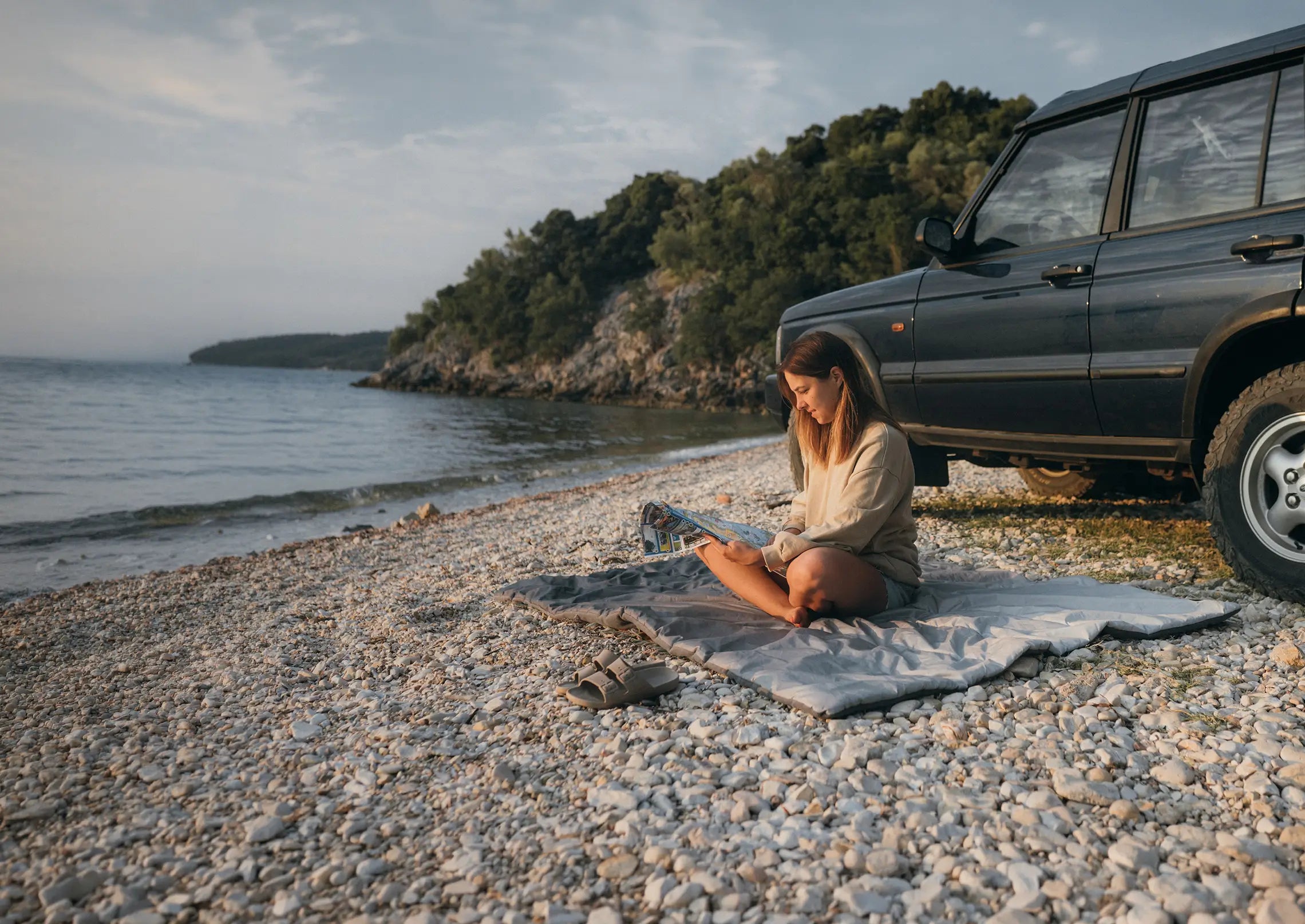 A woman sitting on the Horizon Overland Blanket near a body of water next to her car. 