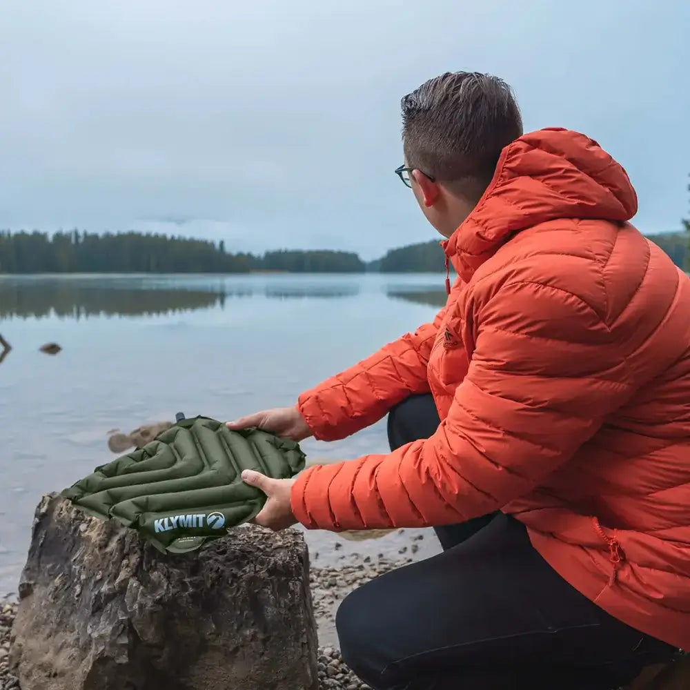 Person in an orange jacket setting up a green Klymit V Seat pad by a lake.