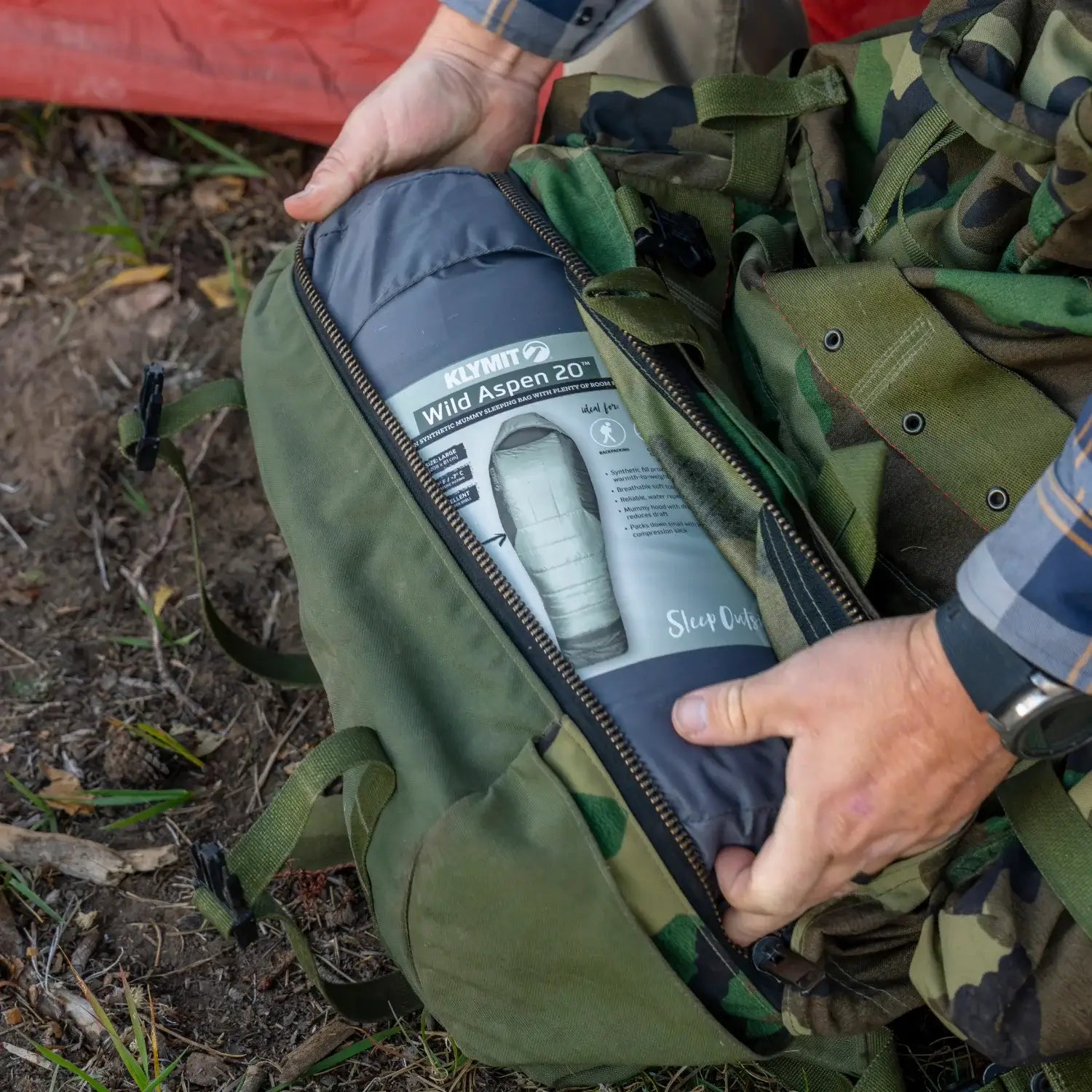 Man pulling a Klymit Wild Aspen 20 Large sleeping bag in its packaging from a backpack at a campsite.