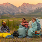 all-groups Three campers in Klymit Wild Aspen sleeping bags at sunset with a mountain backdrop.