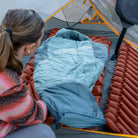 Woman setting up a Klymit Wild Aspen 20 Regular sleeping bag on a Klymit sleeping pad inside a tent, overhead angle.