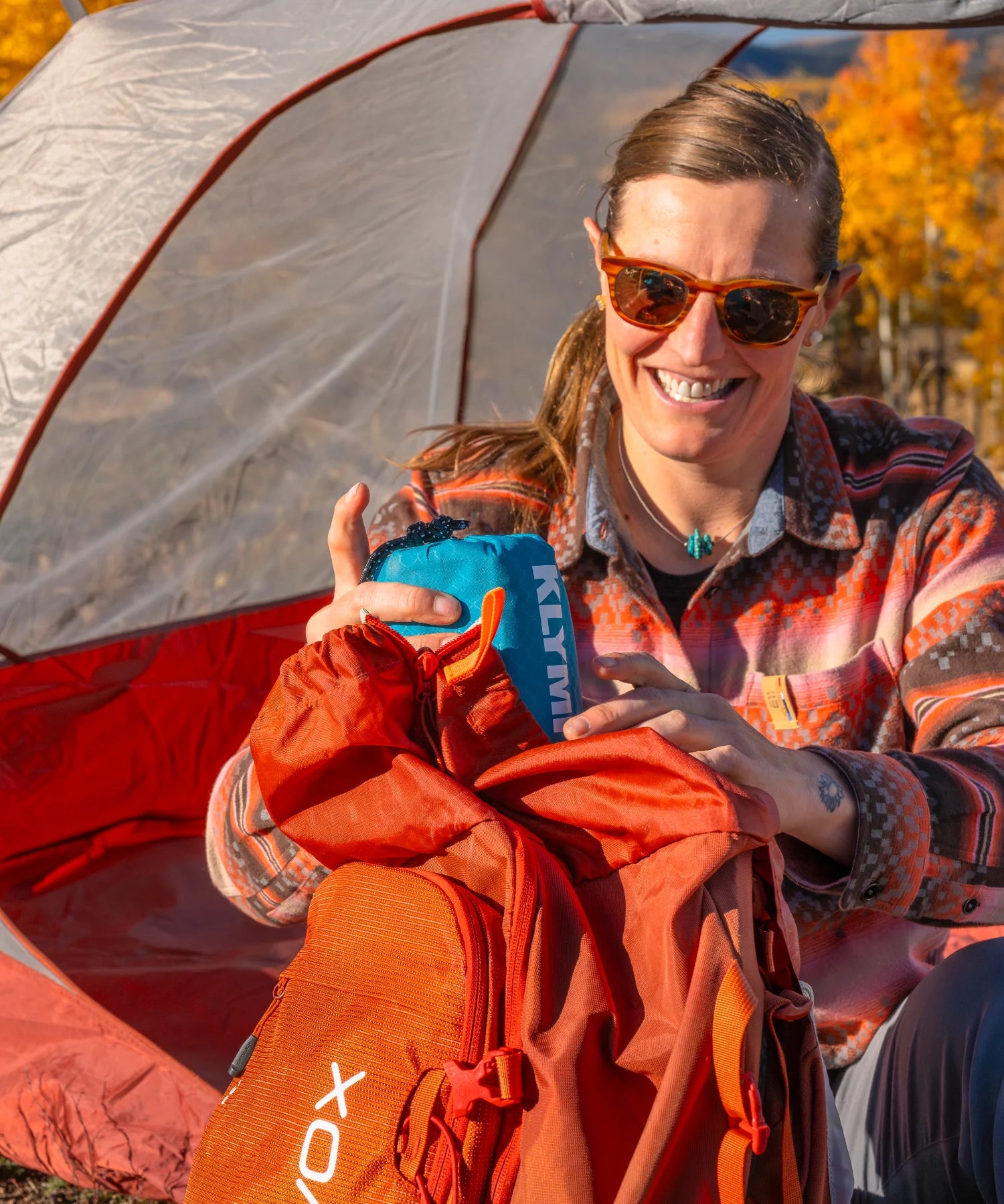 Smiling woman pulling a Klymit sleeping pad stuff sack out of her backpack outside a tent surrounded by golden autumn aspens, featured in the Backpacking collection.