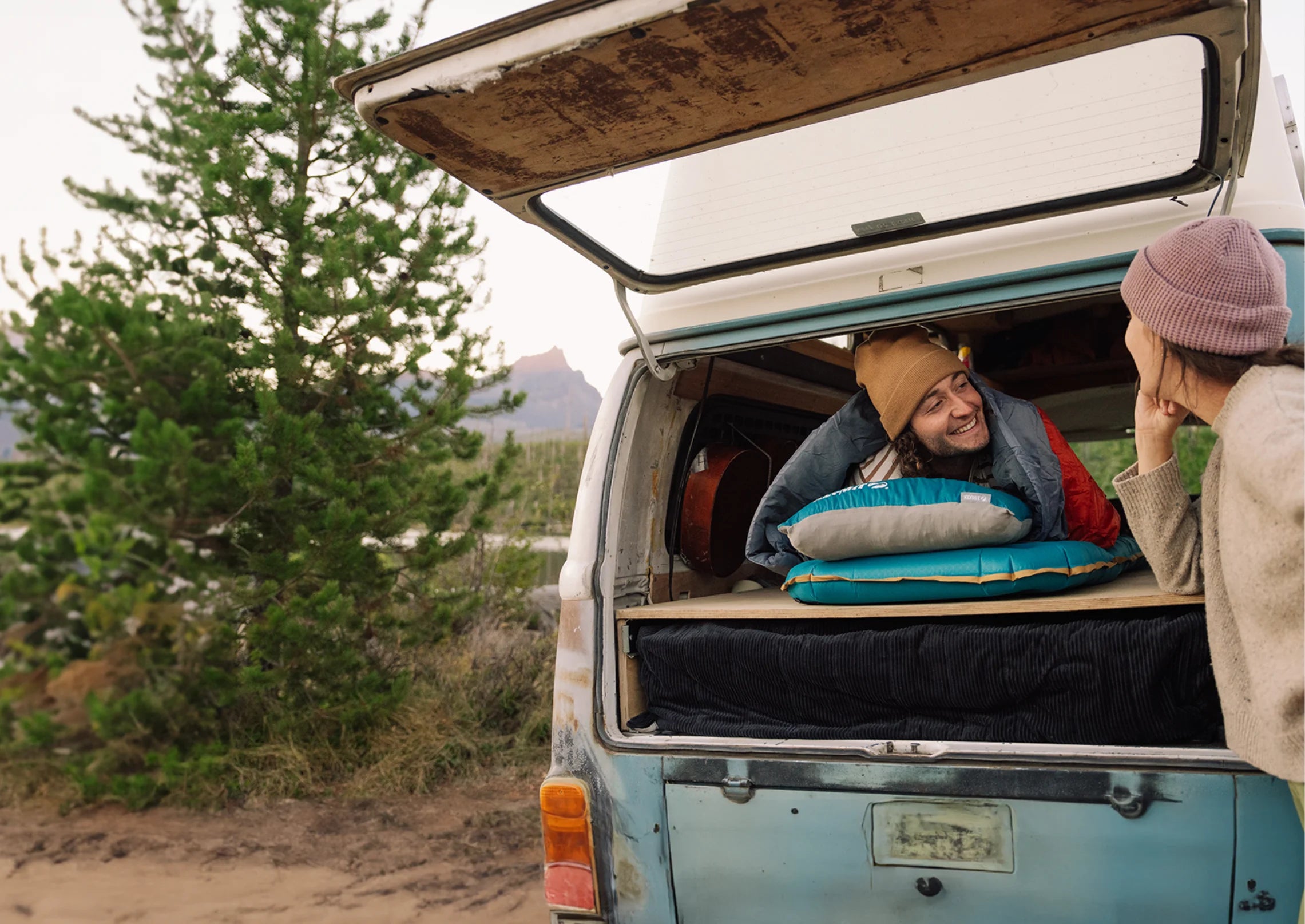 Smiling man lying in the back of a vintage camper van, chatting with a woman outside, featuring a sleeping pad and pillow from the Klymit Base series.