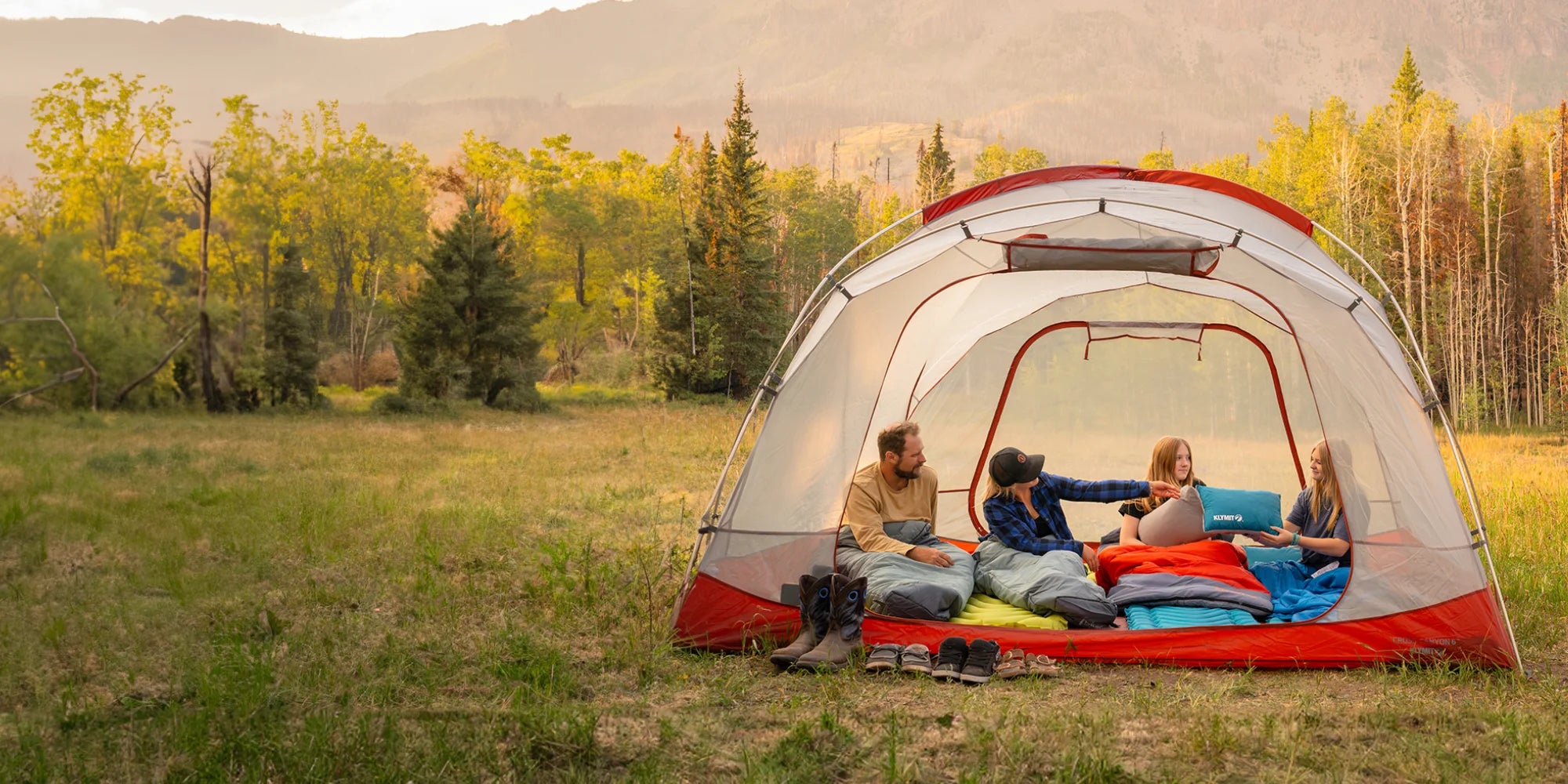 A family sits inside a tent on a lineup of Klymit sleeping pads, sleeping bags, and pillows.