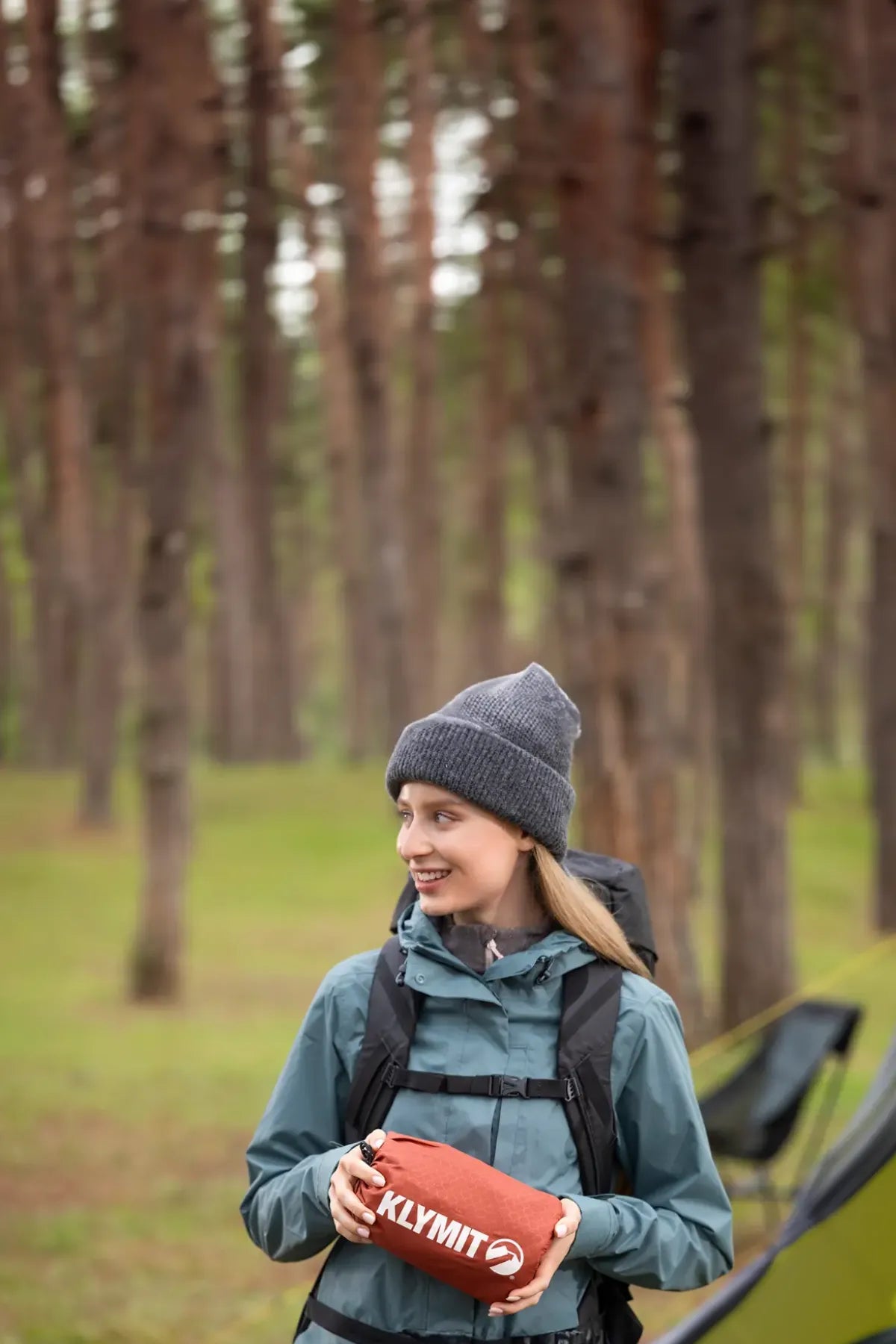 Woman in a beanie and rain jacket holding a red rock-colored Klymit sleeping pad packed in its stuff sack beside a tent.
