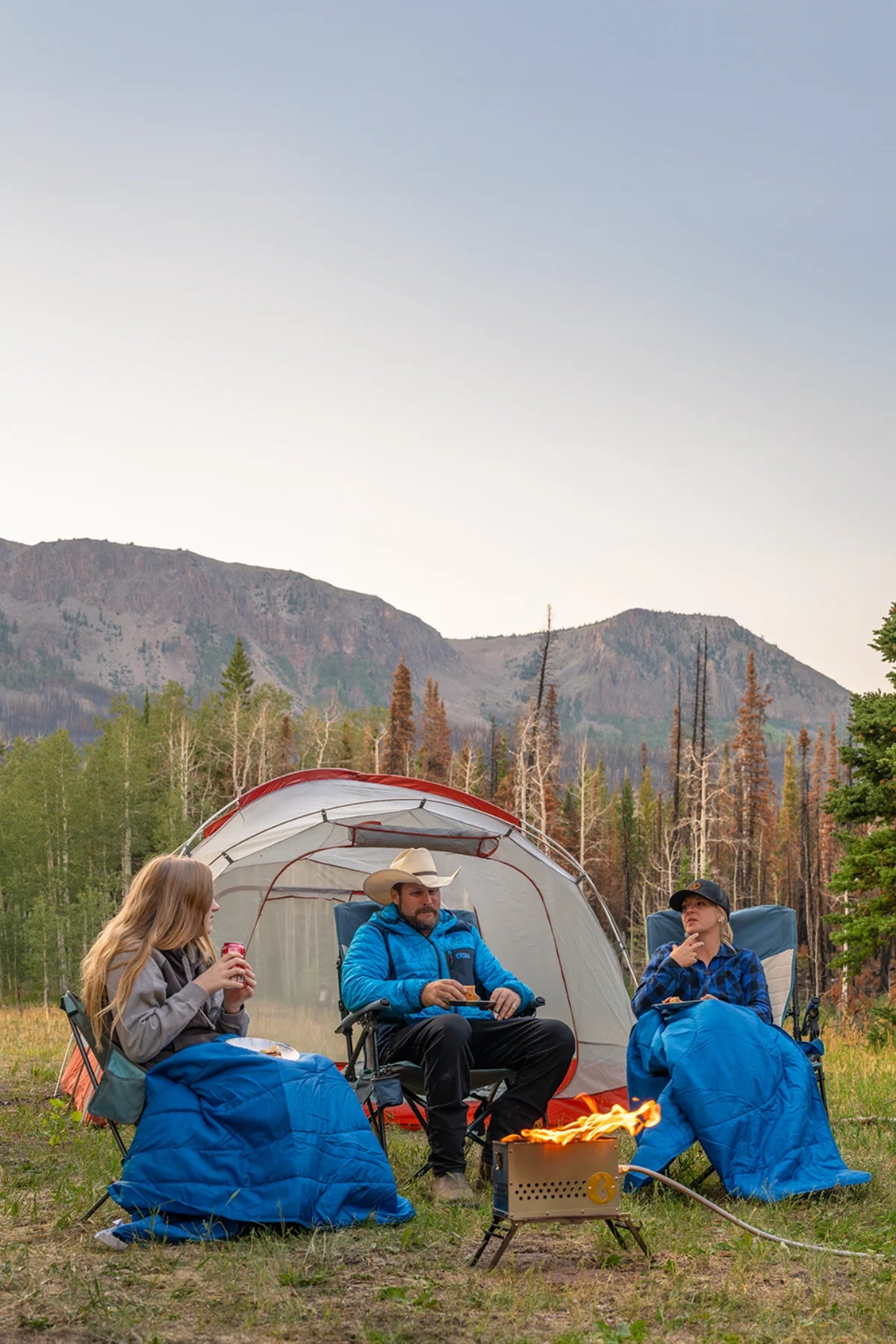 Four people relax around a campfire outside a tent, wrapped in blue Horizon travel blankets, in a mountain meadow at dusk.