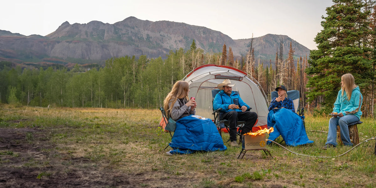 Four people relax around a campfire outside a tent, wrapped in blue Horizon travel blankets, in a mountain meadow at dusk.