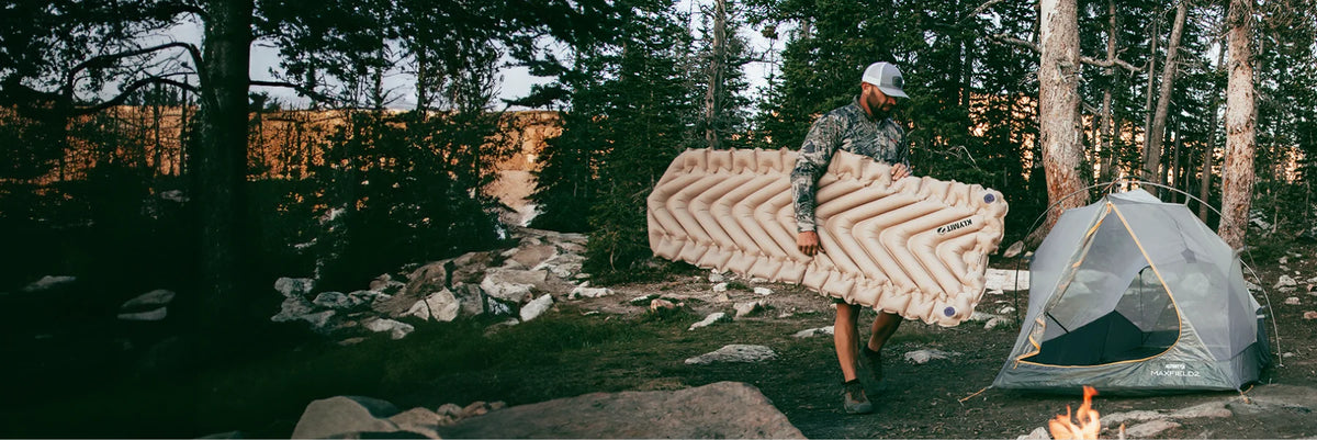 Man carrying a fully inflated Insulated Static V Luxe SL sleeping pad toward a Klymit tent at a rocky forest campsite.