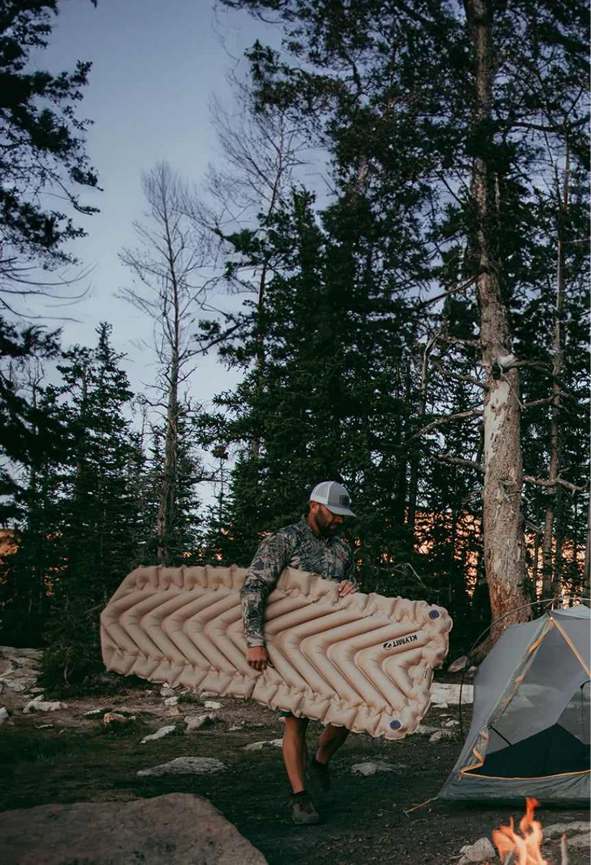 Man carrying a fully inflated Insulated Static V Luxe SL sleeping pad toward a Klymit tent at a rocky forest campsite.
