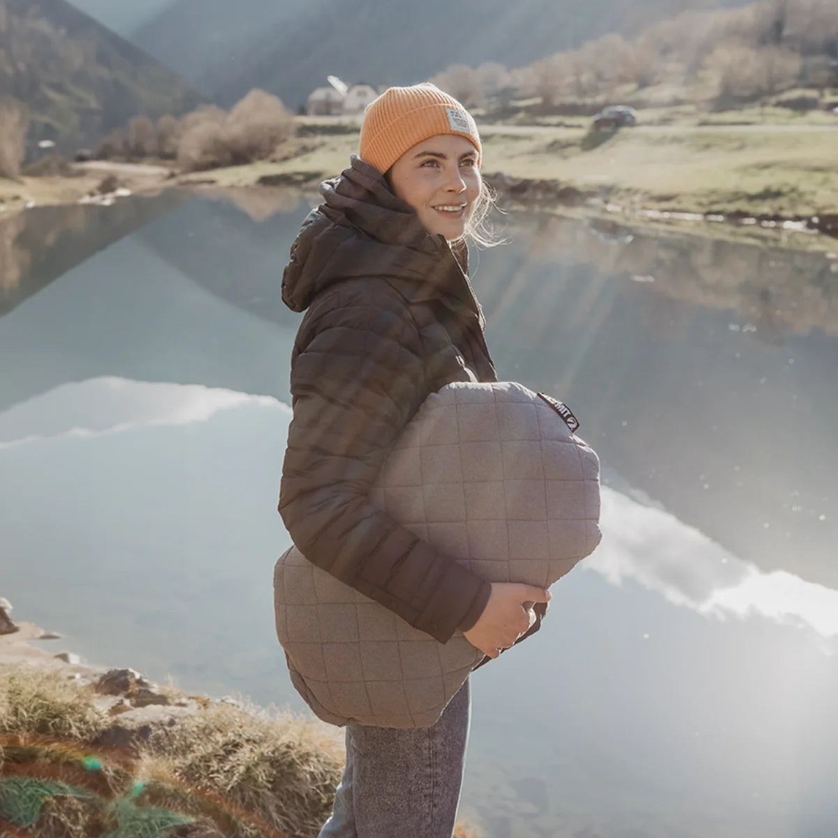Woman carries a gray quilted Pillow X Luxe Summit while standing by a lake in the mountains.