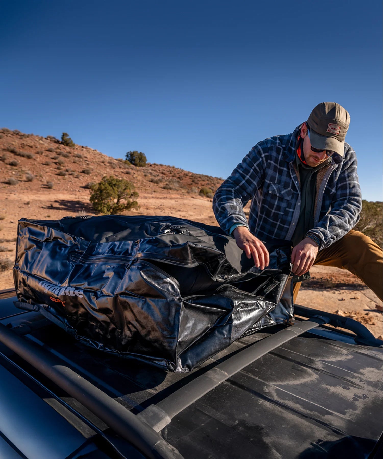 Man loading a Rightline gear waterproof rooftop cargo carrier onto a vehicle roof rack at a remote desert overlanding site.
